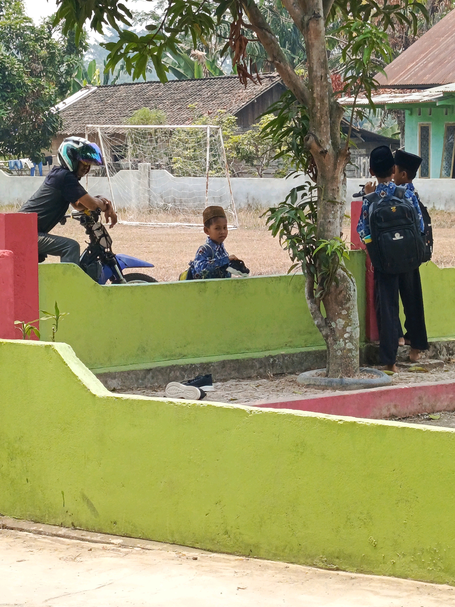 In the city of Megang Sakti V, Indonesia, on a bright day, a man in a black t-shirt and helmet is leaning on a blue motorcycle, appearing to be resting or waiting. Nearby, a young boy wearing a peci and a blue patterned shirt sits on the ground. Two other young boys, also wearing pecis and backpacks, stand near a tree. The background shows a soccer field with a goal, houses with tiled roofs, and lush green trees. The foreground is dominated by a bright green, low wall, with a pair of black sneakers placed on top of it. The scene appears to be an outdoor area, possibly a schoolyard or a community space.