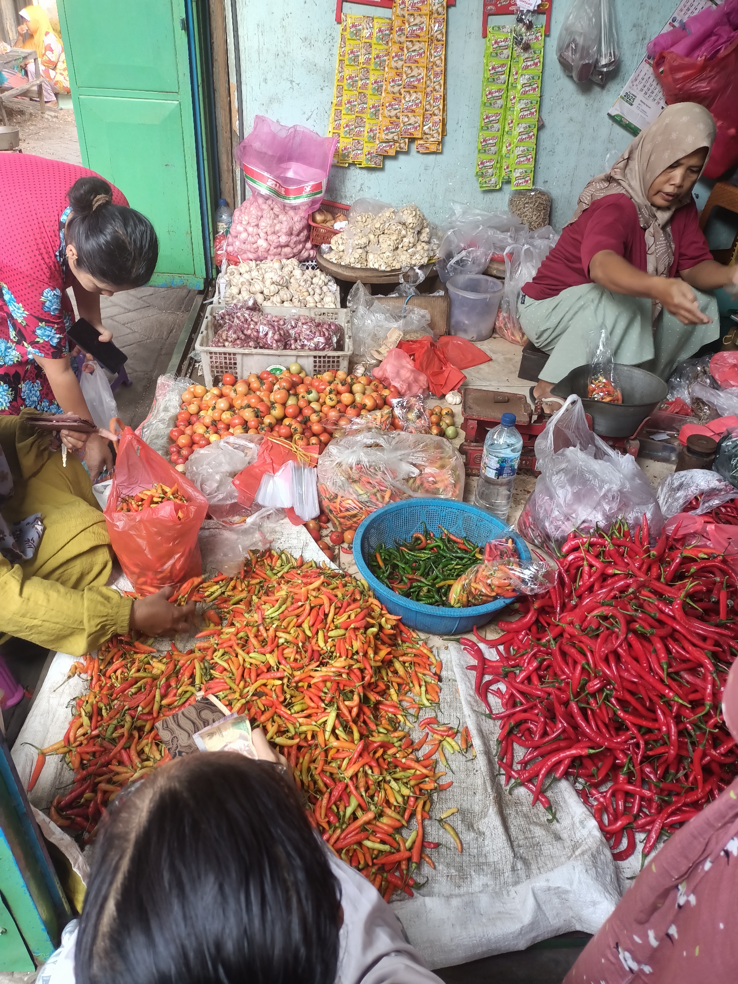 In the city of Badas, Indonesia, a market scene unfolds with an abundance of fresh produce. Several individuals are engaged in transactions or preparing goods. In the foreground, a woman in a yellow top is seen sorting through a pile of red and yellow chilies, her hands actively separating them. To her right, another woman in a maroon top and hijab is crouched, appearing to be selling items or tending to a bin of red chilies. Behind them, a customer wearing a pink and blue patterned top is leaning over, examining items and holding a mobile phone. The market stall is laden with various vegetables, including large quantities of tomatoes, onions, and numerous bags of different types of chilies. Other items visible are garlic bulbs, dried anchovies, and a bottle of water. The background shows a market structure with a green door and walls adorned with colorful packets of seasonings, suggesting a bustling commercial environment. The overall impression is one of a vibrant local market focused on agricultural products.