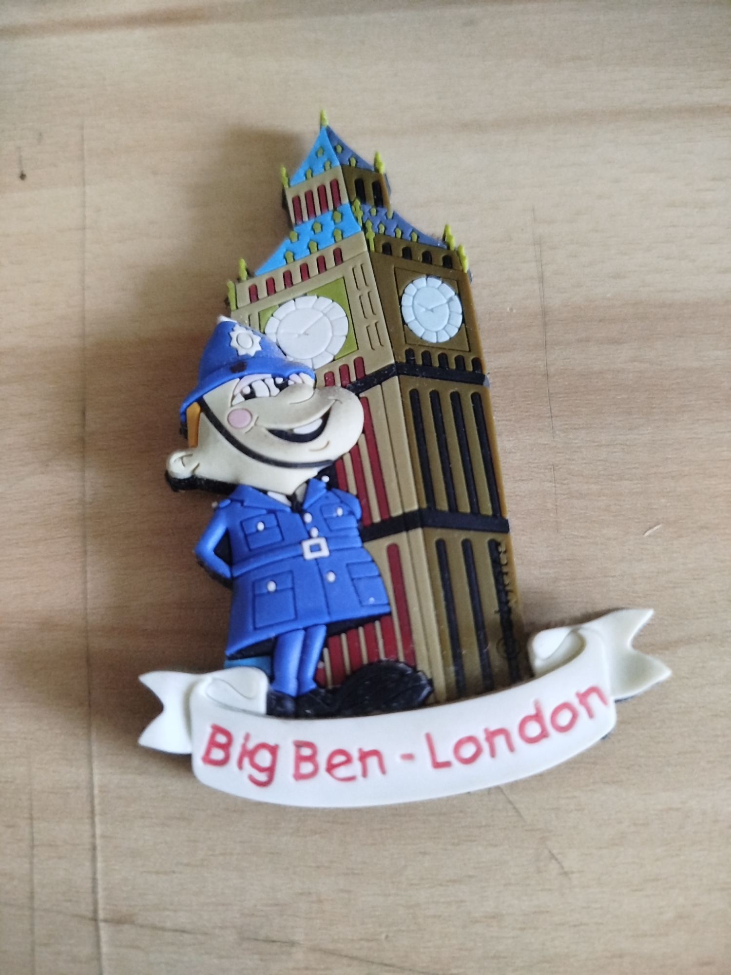 The image shows a souvenir magnet of a cartoon police officer standing in front of Big Ben. The magnet has a white banner at the bottom that reads "Big Ben - London" in red lettering. The police officer is wearing a blue uniform and hat. Big Ben is depicted in brown, blue, and gold colors. The magnet is sitting on a light brown wooden surface. The location in the city of Varna, Bulgaria is not depicted in the image.