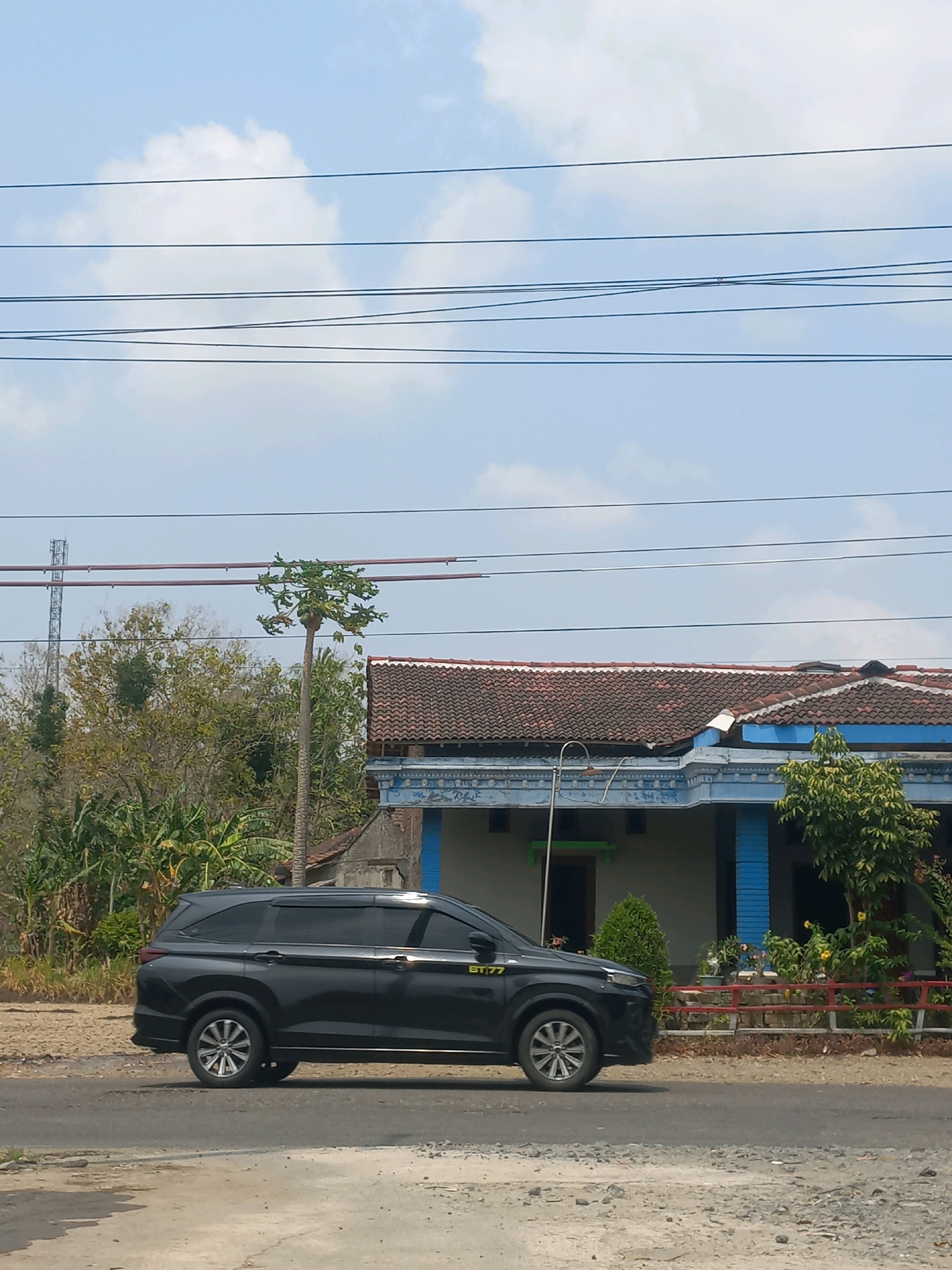 In Baturetno, Indonesia, a black SUV is parked on a dusty road adjacent to a residential building. The building has a red tiled roof and blue trim. Surrounding the property are trees and vegetation, with a red-railed fence in front of the house. Power lines are visible crisscrossing the sky, with a cellular tower in the distance. The sky is clear with scattered clouds.