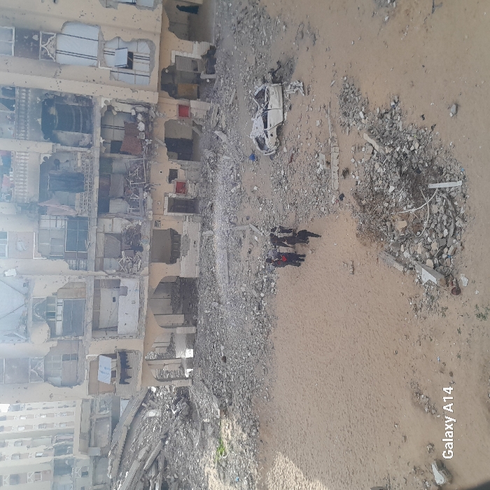 The image displays an outdoor scene in Khan Yunis, Palestinian Territory, characterized by widespread structural destruction under daylight conditions. A severely damaged, multi-story building with a light-colored facade dominates the background. This building exhibits extensive external damage, including large sections of missing walls exposing interior rooms, shattered or absent windows, and numerous impact craters or bullet marks across its surface. Visible debris, including clothes, hangs from balconies and exposed interior spaces. To the left of this structure, a massive pile of rubble indicates a completely collapsed building. Other damaged structures are visible further in the distant background.
The ground in the midground and foreground is a desolate mix of a central sandy patch surrounded by and interspersed with dense fields of pulverized concrete, twisted rebar, and miscellaneous building debris. Small pools of standing water are discernible at the base of the large damaged building.
In the mid-left portion of the sandy area, a group of approximately six individuals, dressed in dark clothing, are gathered. One individual is slightly separated from the main group and appears to be walking towards the right. To the right of this group, positioned on the rubble, lies the extensively damaged and likely burned chassis of a white vehicle, stripped of tires and most body panels. Various smaller fragments of concrete and metal debris are scattered throughout the scene.