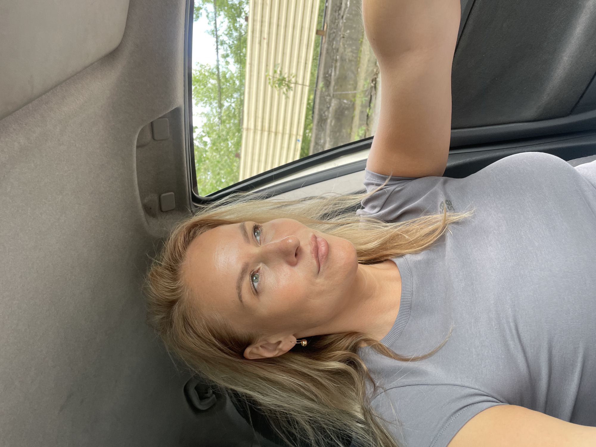 A blonde woman wearing a grey t-shirt is lying in the back of a vehicle. She is positioned with her head near the car's interior side panel and her shoulder resting against the window. Outside the window, a corrugated metal structure and greenery are visible. The location is Baranovka, Russia.