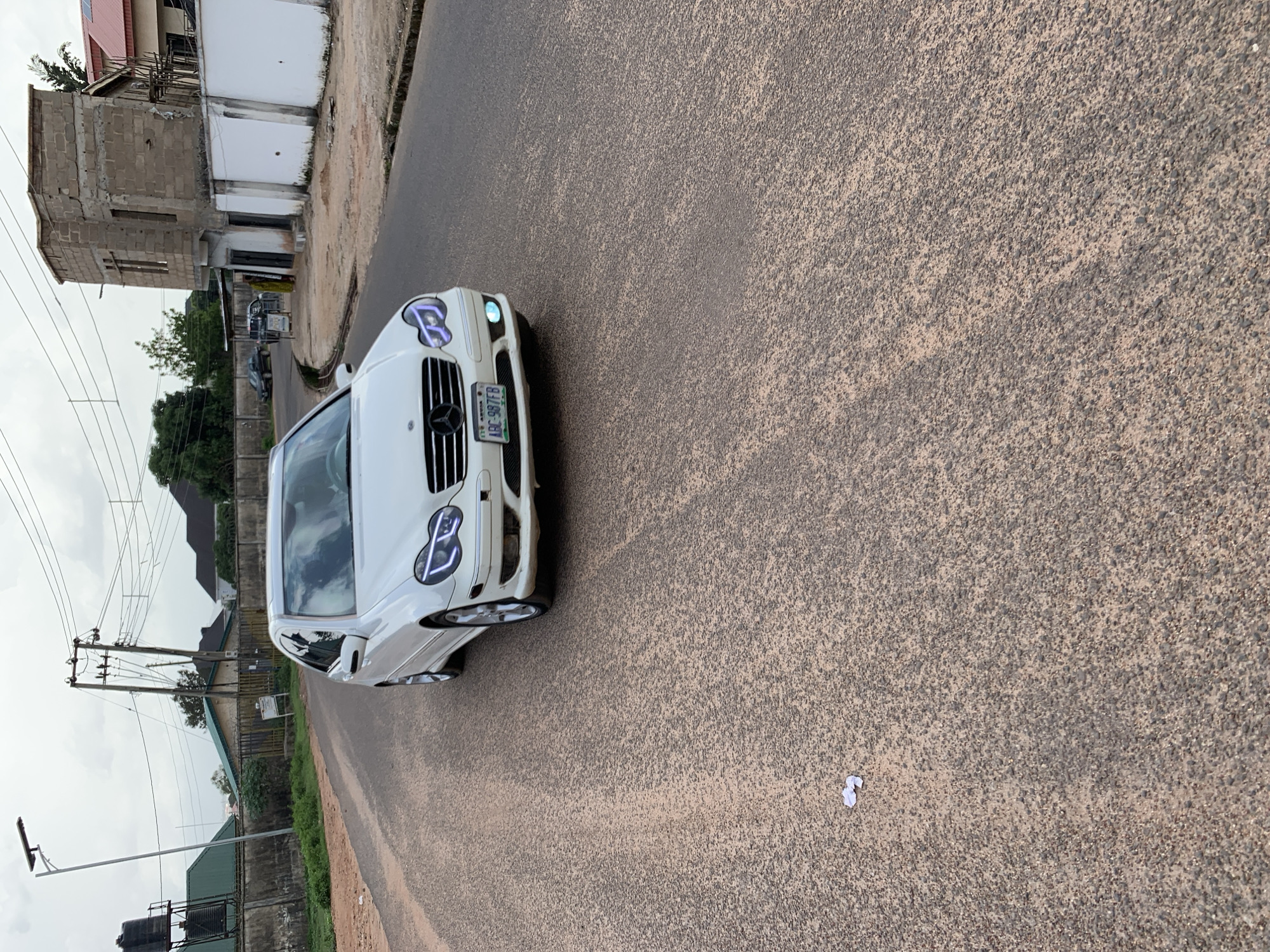 A white Mercedes-Benz C-Class sedan with license plate ABC-987FB is parked on a paved road in Jos, Nigeria. The car is facing the camera and its headlights are on, with a distinctive LED strip. The road is rough and appears to be made of asphalt with gravel. In the background, there are several buildings, including a partially constructed structure made of cinder blocks and a white-walled building. Power lines and a solar-powered street light are also visible. A few scattered pieces of white paper are on the road in the foreground. The sky is overcast, suggesting a cloudy day. The image is taken from a low angle, emphasizing the car and the road.