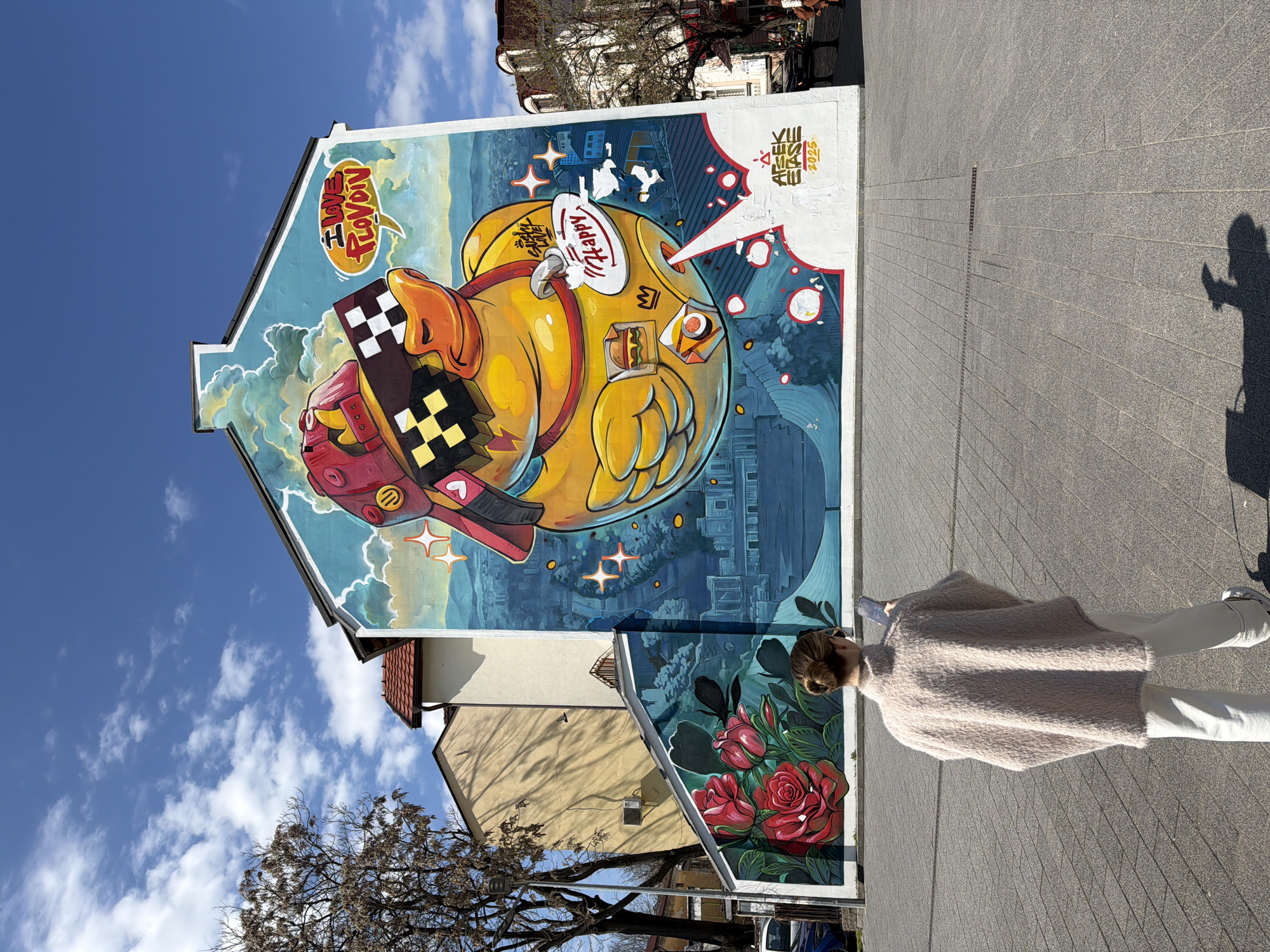 The image depicts a large mural on the side of a building. The mural features a cartoon duck wearing sunglasses and a sneaker, with city elements incorporated into the background. The mural also includes text "I LOVE PLOVDIV". At the bottom of the mural are red roses. A person stands in front of the mural, dressed in white pants and a light pink sweater. They have their hair in a bun. The person is facing away from the camera. The image also includes a view of the sky with some clouds, and other buildings are visible. The image is taken in Plovdiv, Bulgaria, due to the inscription "I LOVE PLOVDIV".