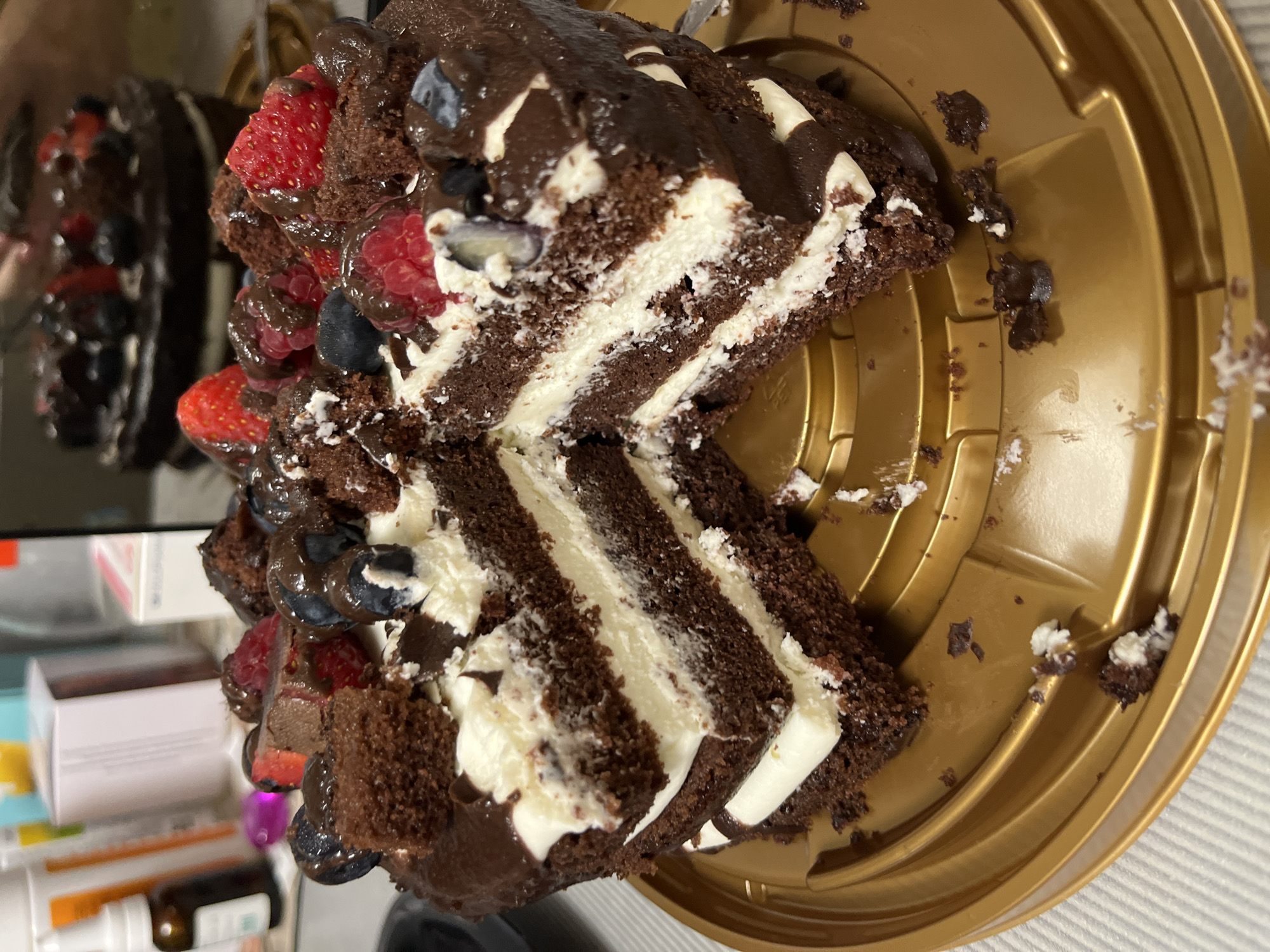 A rich chocolate cake, layered with white cream and topped with fresh berries and chocolate ganache, is presented on a golden serving platter. The cake has been sliced, revealing its intricate structure. Crumbs and a small fork are visible on the platter. In the background, out-of-focus elements suggest an indoor setting, possibly a kitchen or dining area, with various objects on shelves, including bottles and boxes. The image does not contain any elements that would allow for the determination of a location in Sochi, Russia.
