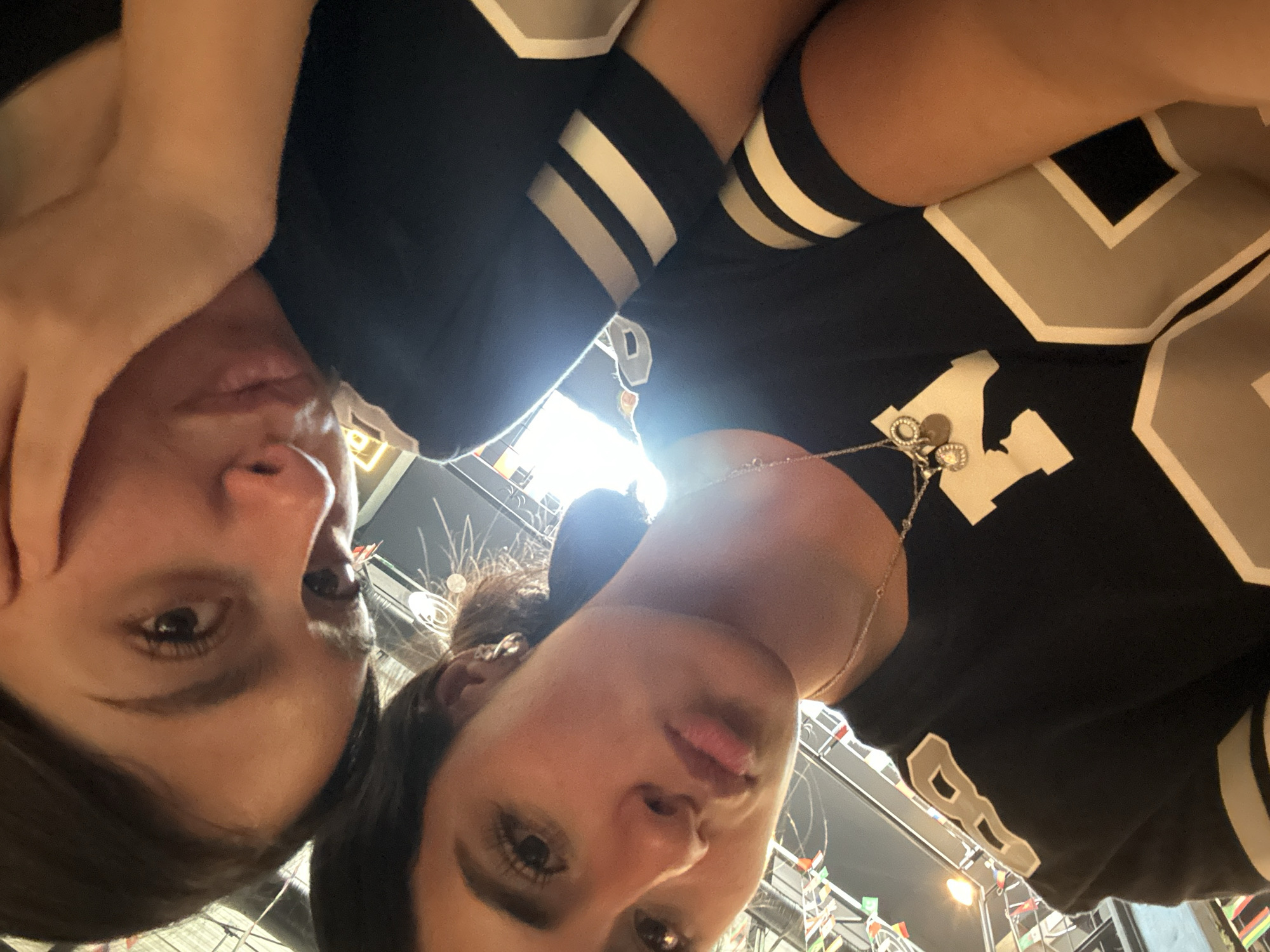 Two women are close together in a selfie, oriented from a low angle, capturing their faces and upper bodies. One woman on the right wears a black jersey with large, light-colored numbers and stripes on the sleeves. She has a necklace with multiple charms. The second woman is on the left, her hand near her face. The background reveals an interior space with flags and lighting fixtures. The specific location is not identifiable.