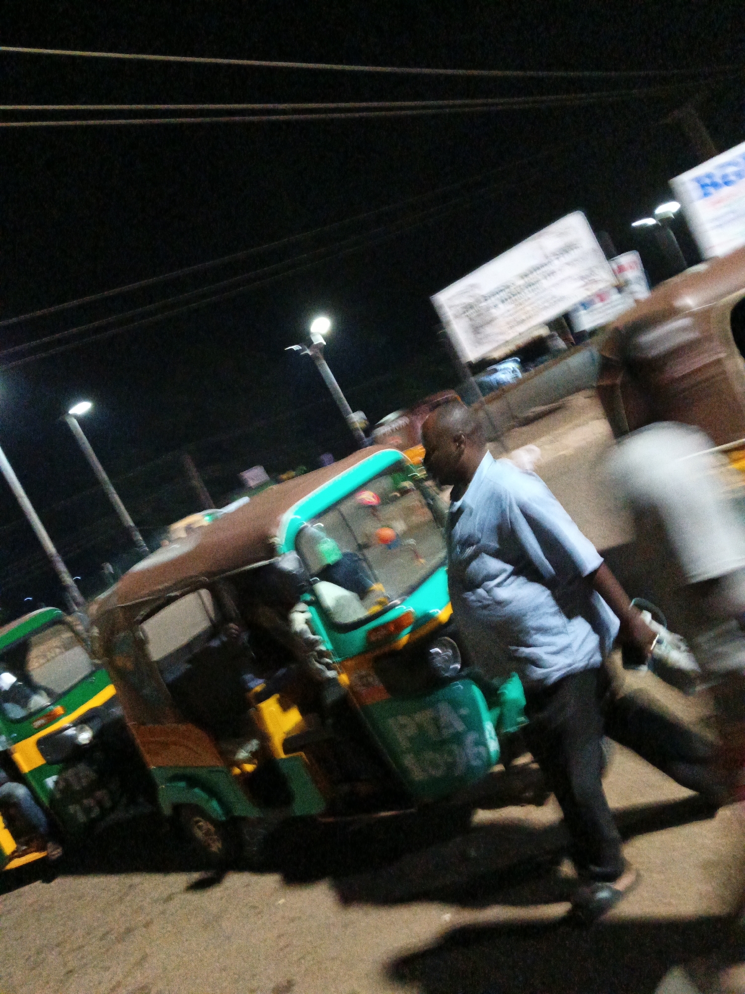 In Bukuru, Nigeria, at night, a person wearing a light blue collared shirt and dark pants walks near several tuk-tuks. The tuk-tuks are green and brown with some yellow accents. One tuk-tuk has "PTA-1036" written on its side. The scene is illuminated by streetlights, casting long shadows. Several signs are visible in the background, one with blue and white text. The ground appears to be a paved surface. The image has a motion blur effect, indicating movement.