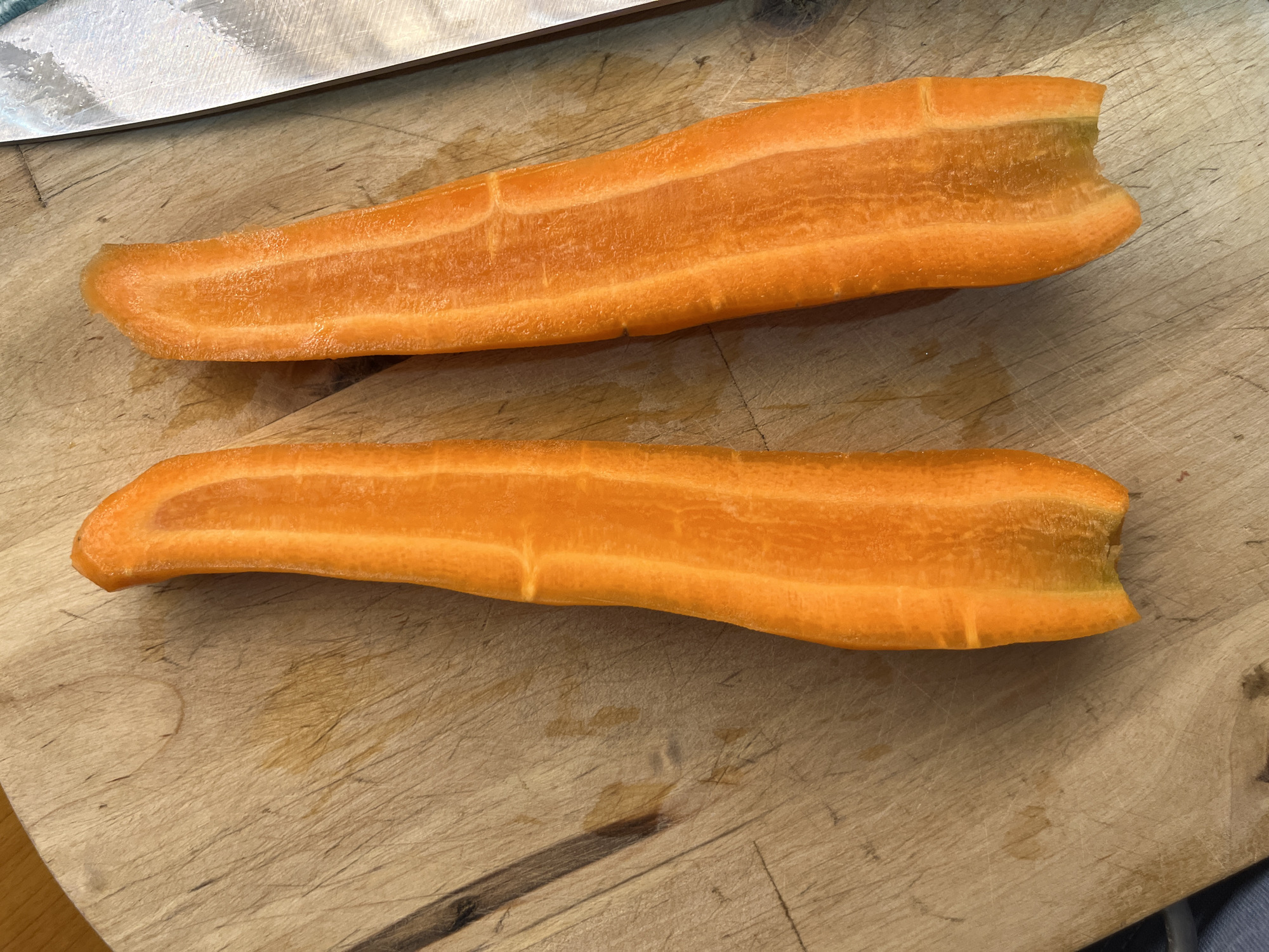 Two halves of a peeled and cut carrot lie on a wooden cutting board. A portion of a knife is visible on the right side of the image. The cutting board has visible scratch marks and some orange stains. The carrots are a bright orange color with some lighter streaks visible in their flesh. The scene appears to be indoors, likely in a kitchen setting, based on the presence of a cutting board and knife. No people or other objects are visible.