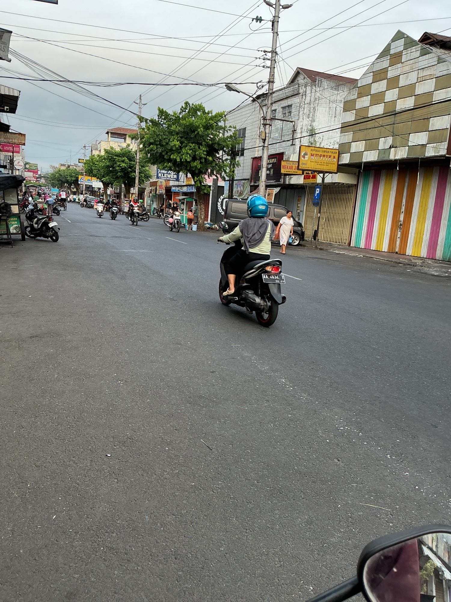 The image depicts a street scene in Magelang, Indonesia, likely during the daytime with overcast skies. A person wearing a blue helmet and a light-colored top over black pants is riding a dark-colored scooter in the foreground, traveling away from the viewer. To the left of the scooter rider, a woman in a light-colored dress is walking on the sidewalk. The street is paved with asphalt and is lined with buildings on both sides.
On the right side of the street, several shops are visible. One building has a distinctive checkered facade with green and beige squares. Adjacent to it are roll-up doors painted with vertical stripes of pink, yellow, orange, and blue. A yellow sign with text in Indonesian is mounted on a pole in front of another building.
On the left side of the street, more buildings house various businesses, indicated by signs. A prominent sign reads "TOKO KELLER". A variety of motorbikes are parked along the sidewalk.
Further down the street, in the background, more buildings and trees are visible, along with a multitude of other motorbikes and some pedestrians. Overhead, a complex network of power lines crisscrosses the sky. The overall impression is of a busy urban street in Indonesia.