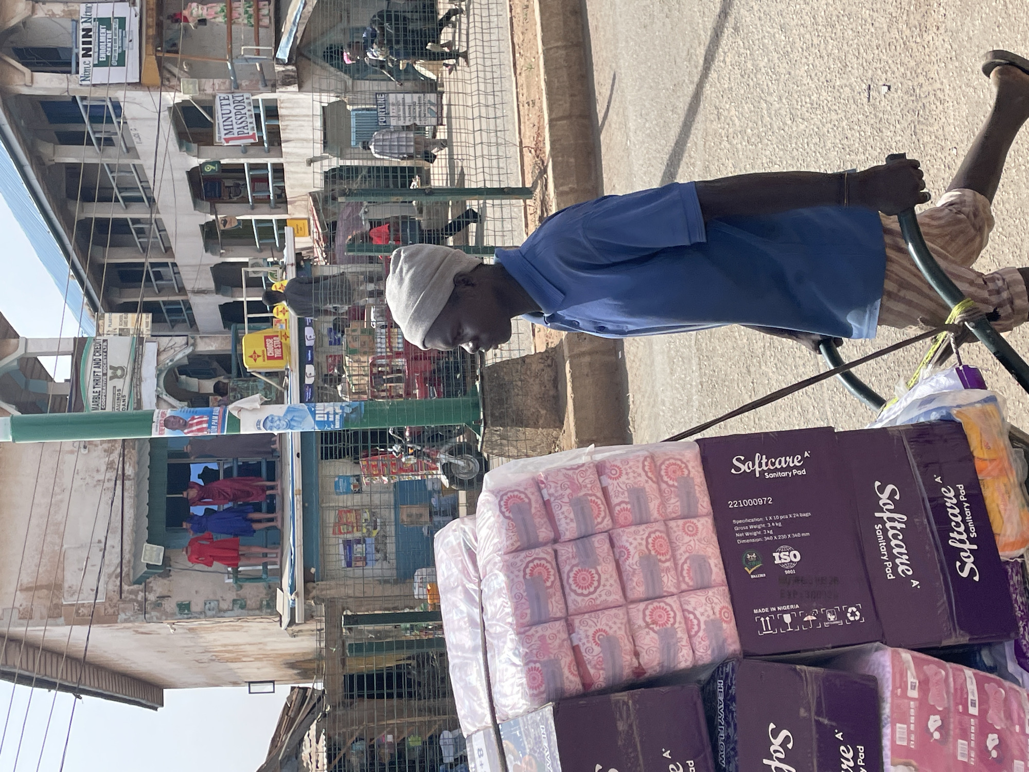 The image captures a vibrant street scene in Bukuru, Nigeria, under clear, bright daylight. In the foreground, a man with dark skin, wearing a blue polo shirt and a light-colored head covering, pushes a metal cart laden with merchandise. He is seen in profile, his hands gripping the cart's frame, and he wears sandals on his feet.
The cart is primarily filled with large quantities of "Softcare Sanitary Pad" products. The packages are distinct, with some in purple boxes displaying detailed specifications like "MADE IN NIGERIA," "ISO 9001," "Gross Weight 3.4 kg," and "Dimension: 340 X 230 X 340 mm." Other packages are in pink plastic wrapping, featuring floral patterns and marked "HEAVY FLOW."
The background reveals a bustling urban environment with multi-story buildings lining the street. These structures feature numerous windows and balconies, and some appear to be commercial establishments. Several signs are visible:
* "Nime NIN Nime ENROLMENT CENTRE"
* "1 MINUTE PASSPORT"
* "MARBLE THRIFT AND CREDIT COOPERATIVE SOCIETY LTB." with partial text "AVINGS DANS"
* A yellow sign reading "CHOP THE STAR"
* A sign partially visible with "SCHOOL"
* A banner with faces reading "OR PAM BAS"
* The word "FORTUNE" is visible on a fence.
Other details in the background include a metal fence, glimpses of other individuals, and what appears to be mannequins dressed in red and blue outfits displayed in a storefront window. The street surface is paved, suggesting an active commercial area. The overall scene conveys a sense of everyday commerce and activity in an African city.