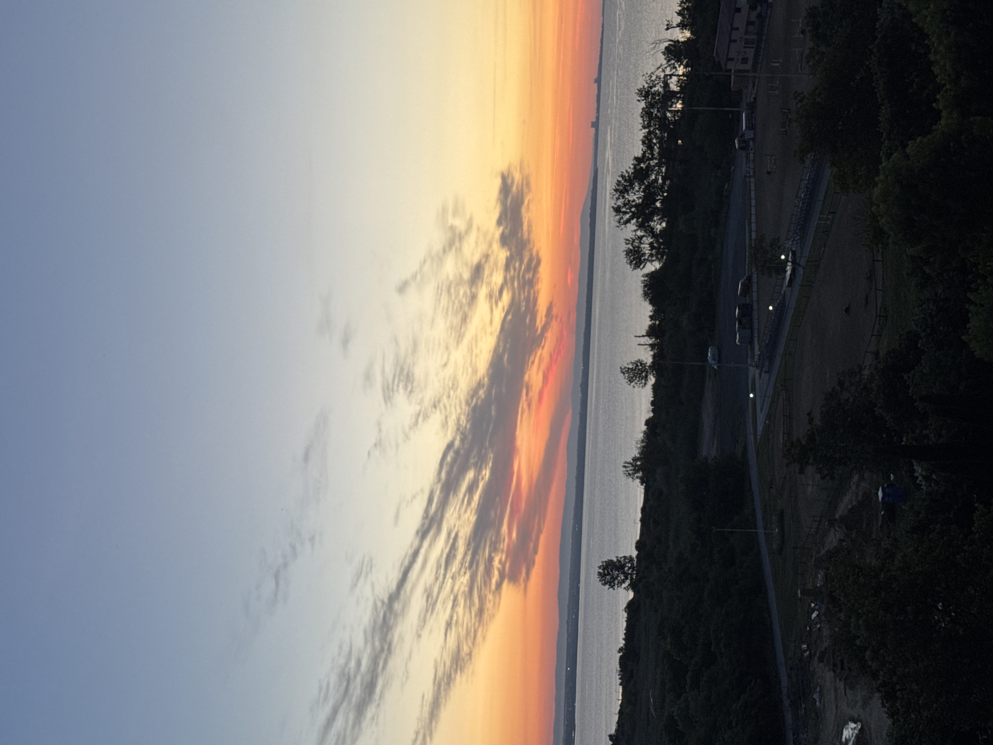 This is an eye-level, wide shot of a body of water at sunset. The sky transitions from a light blue to orange and pink, with wispy clouds. In the distance, a faint, mountainous horizon is visible, possibly separating land from sky. The water is calm and reflects the colors of the sunset. In the foreground, a dark, silhouetted landscape of trees and vegetation is present. Below the treeline, there appears to be a developed area with structures and open spaces, possibly a park or recreational facility. There are no visible people in the image. The overall impression is a tranquil, natural scene at dusk. The location is the Burgas Bay, part of the Black Sea, near the city of Burgas, Bulgaria.