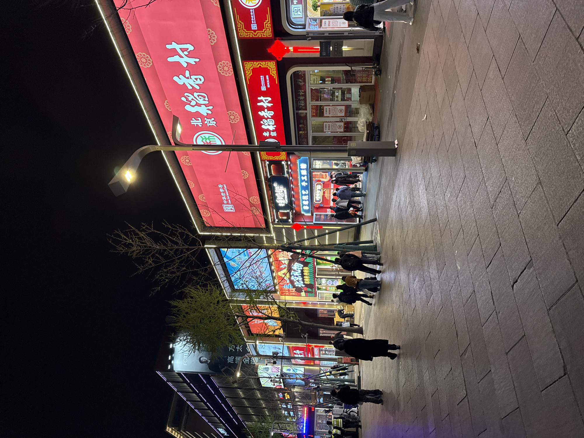 The image shows a street scene at night in what appears to be Dongcheng District, China. Brightly lit shops with prominent Chinese characters are visible. People are walking along the sidewalk. A street lamp illuminates the scene. The businesses appear to be a mixture of traditional shops and potentially modern establishments.