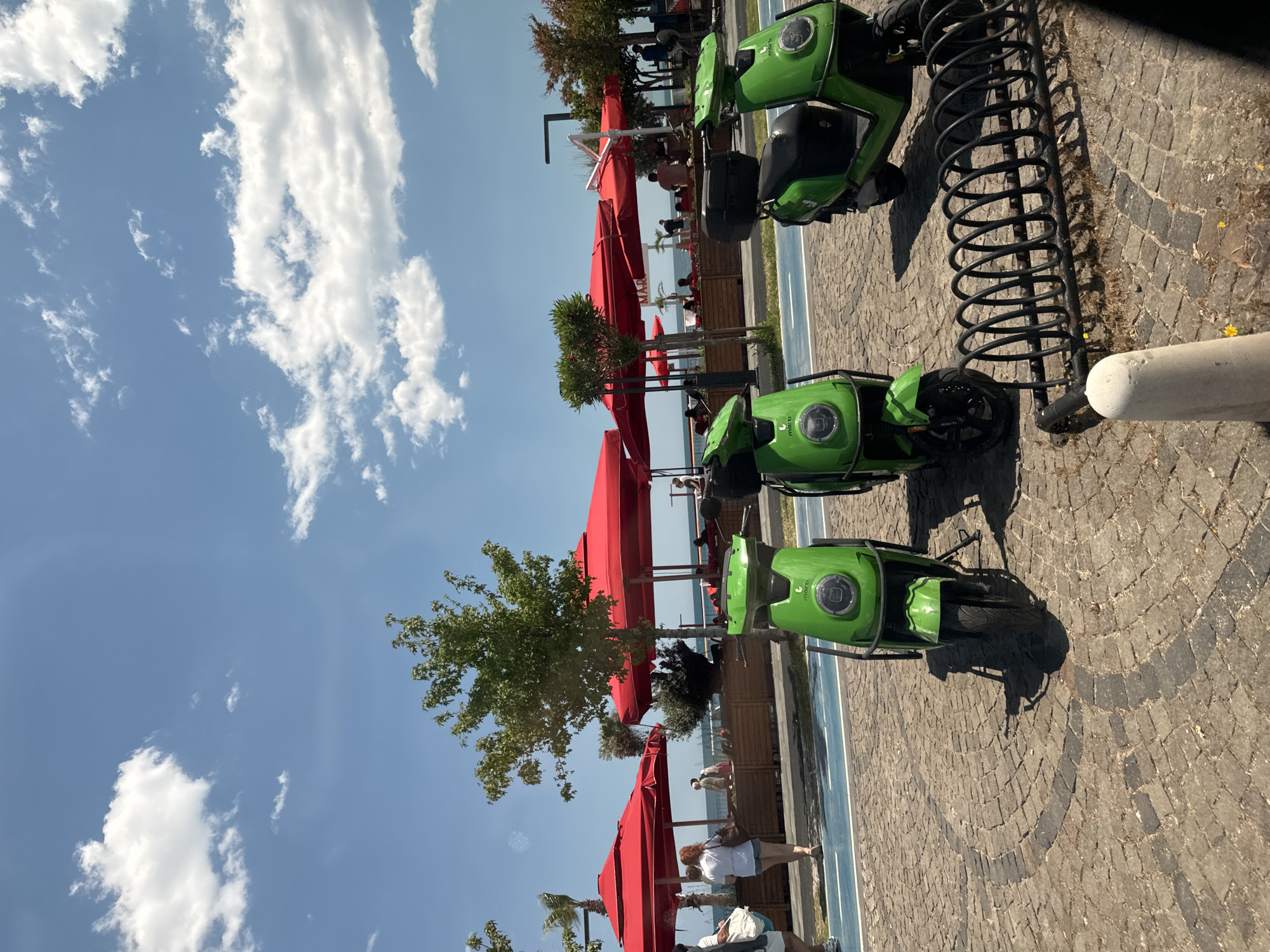The image shows a row of green electric scooters parked along a seaside promenade in Konyaaltı, Turkey. Red parasols shade outdoor seating areas of cafes or restaurants. People are seen seated under the parasols, potentially patrons enjoying the sea view. Beyond the seating area, the blue sea meets the horizon. The sky is blue with scattered white clouds. A distinctive spiral bike rack is visible near the scooters. The promenade is paved with cobblestones. A blue-painted bike lane runs alongside the scooters and outdoor seating.