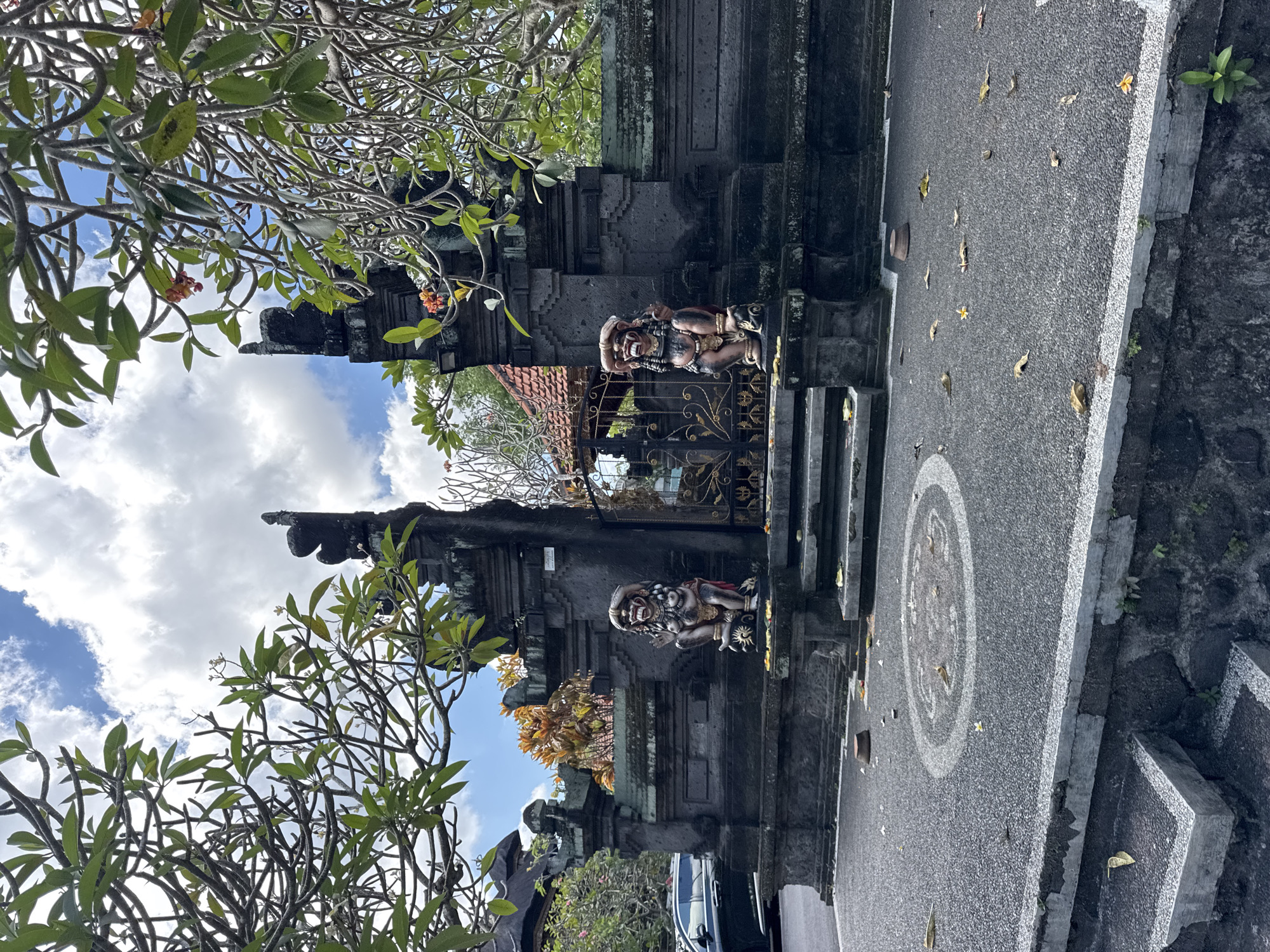 This image captures an ornate Balinese entrance gate, likely leading to a temple or a traditional compound, under a bright blue sky with scattered white clouds. The scene is lush with vegetation, dominated by several frangipani trees (Plumeria) whose gnarled branches, green leaves, and some reddish-orange and white flowers frame the sky and parts of the gate.
The central feature is a dark, intricately carved stone gate, characteristic of Balinese architecture. It features multiple tiers and steps. Flanking the golden, decorative metal gate are two colorful guardian statues, likely mythical Rakshasa or Bhoma figures. These statues are depicted with fierce expressions, fanged mouths, wide eyes, and elaborate headwear, rendered in various earthy and vibrant tones. One statue has a raised hand, and both appear to be in protective poses. A small, unreadable white sign is affixed to the stone structure above the right-hand statue.
Behind the metal gate, a traditional building with a reddish-brown tiled roof is partially visible. The paved ground in front of the gate features a large circular pattern, possibly a mandala or geometric design, inlaid in lighter concrete or paint. Scattered fallen leaves and frangipani blossoms are visible on the grey pavement. A silver car is partially seen on the far right, behind some foliage. The clear light suggests it is daytime, and the overall scene evokes the cultural richness and traditional craftsmanship found in North Kuta, Indonesia. No people are visible, and no specific activity is taking place.