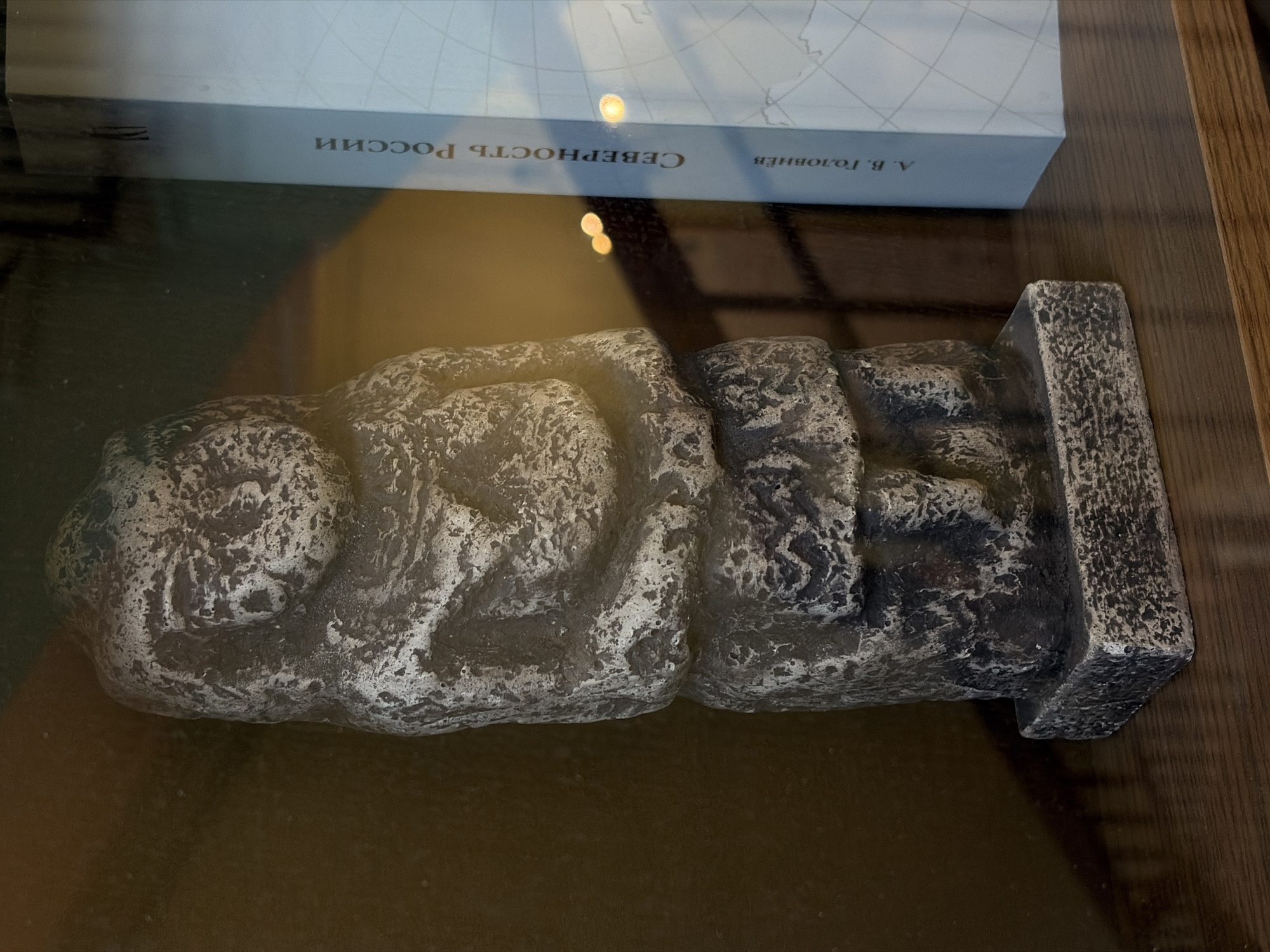 The image shows a rock-like sculpture displayed in a glass case, likely in a museum or exhibition. To the upper-left of the sculpture lies a book with text in Russian, which reads "Северность России" (Severnost Rossii). The name "А. В. Головнёв" (A. V. Golovnev) appears to the right of the title. The sculpture features rough, uneven textures that suggest it is made of stone or a material resembling stone. It has a distinct, rectangular base. The lighting is somewhat dim and diffused. The surface beneath the glass case is a dark, polished wood. Without further context, it is impossible to determine its location within Saint Petersburg.