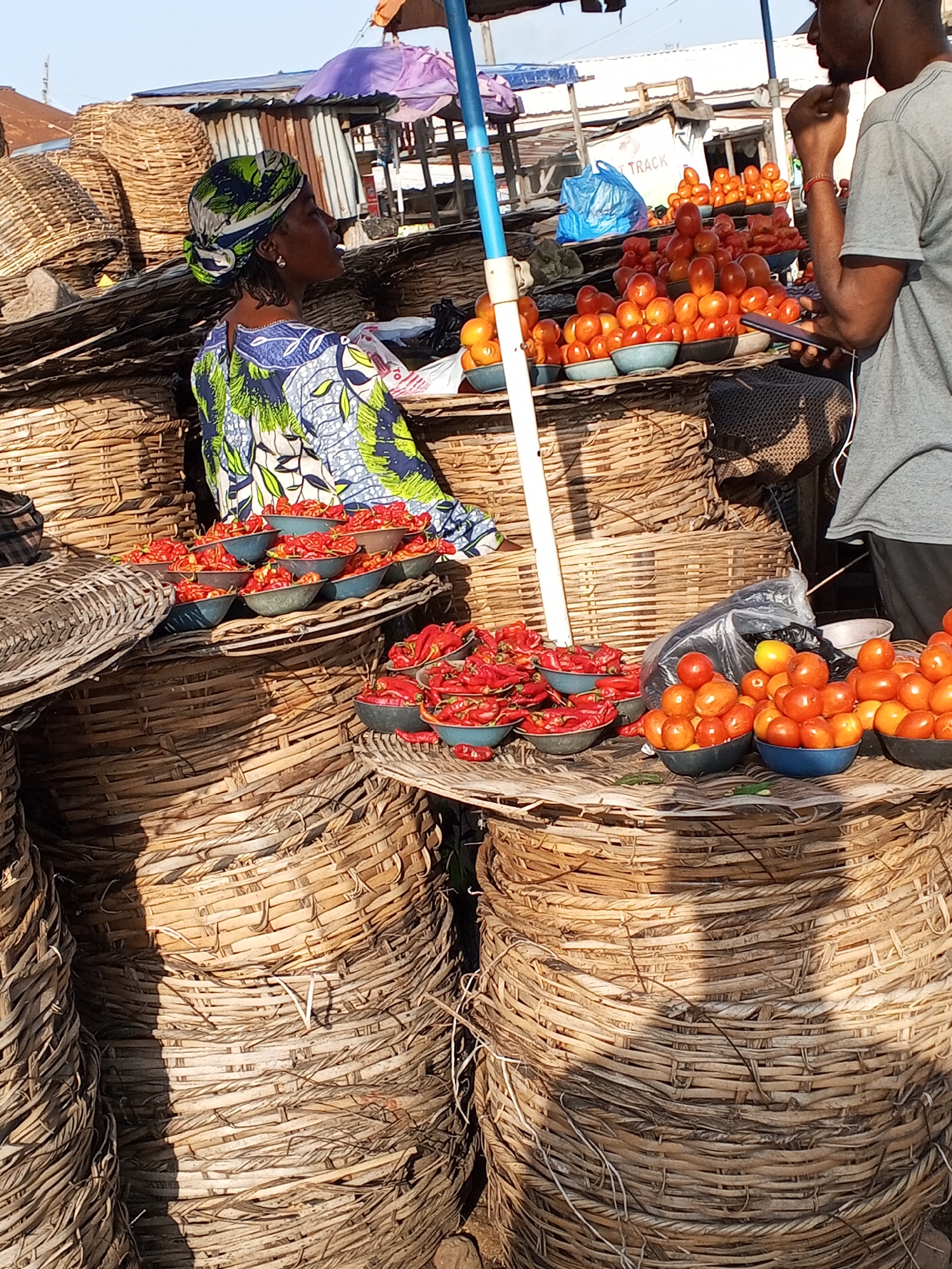 The image depicts a market scene in Lokoja, Nigeria, characterized by an abundance of woven baskets and fresh produce. In the foreground, large, round wicker baskets dominate the scene, some appearing empty and others filled with various items. A woman wearing a green and blue patterned headwrap and a blue and green patterned garment stands amidst the baskets, looking to her right. In the mid-ground, several bowls of red chili peppers are displayed on woven surfaces. Further back, a man in a grey t-shirt is visible, looking at his phone and holding a hand to his ear, with a stack of baskets behind him. Behind the produce, there are makeshift structures covered with corrugated metal and tarps, suggesting a bustling market environment. The overall impression is one of a vibrant, outdoor marketplace with a focus on agricultural products.