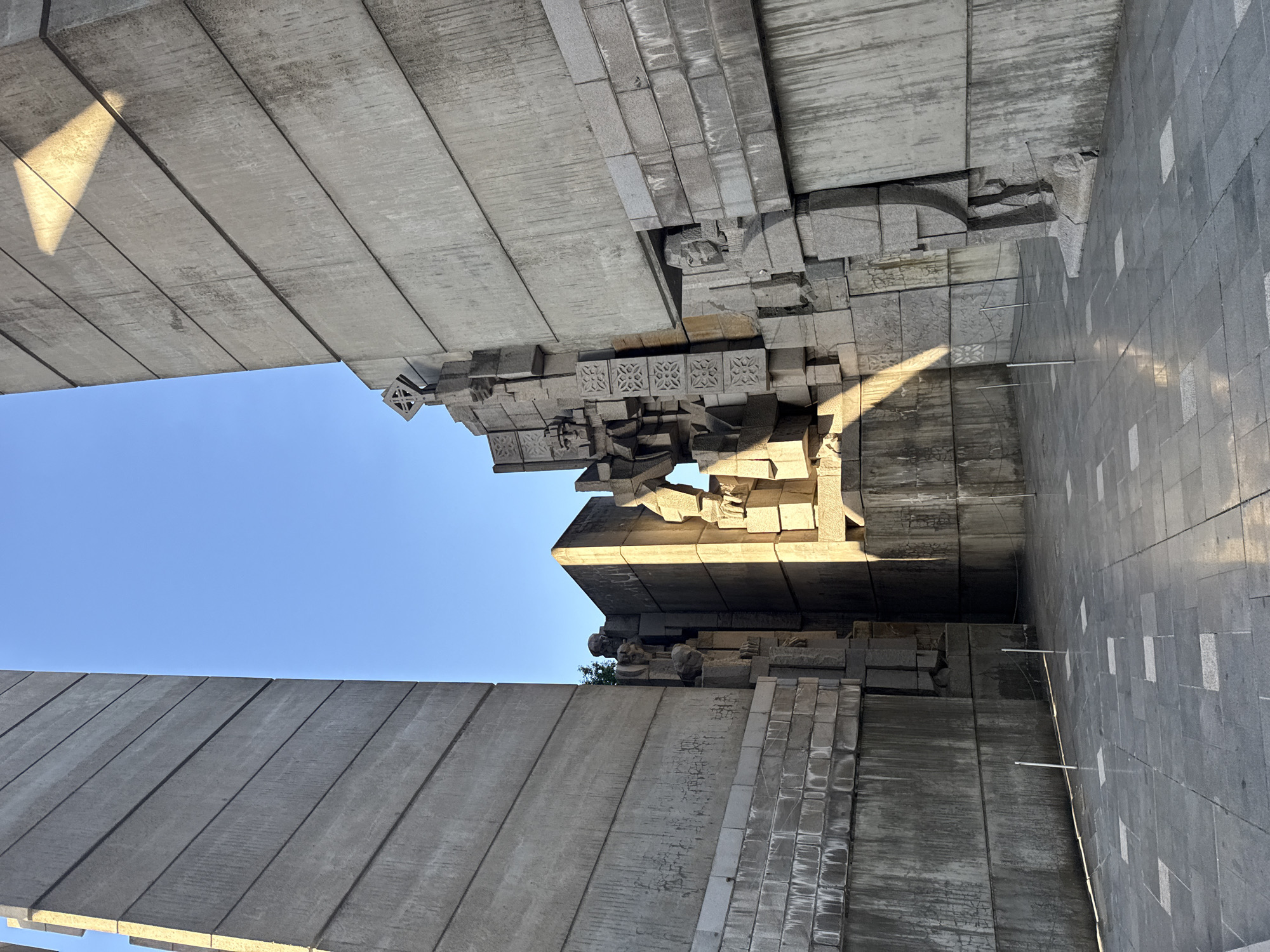 The image depicts a portion of the Founders of the Bulgarian State Monument in Shumen, Bulgaria. The view is angled upwards, showcasing the monument's architectural details and the clear blue sky visible between the concrete structures. The monument is constructed of massive concrete slabs and intricately carved stone blocks. Some of the stone blocks are decorated with geometric patterns. The light suggests it is daytime, with sunlight casting shadows on the stone surfaces. There are no people visible in this shot. The environment consists of the urban landscape of the city of Shumen.