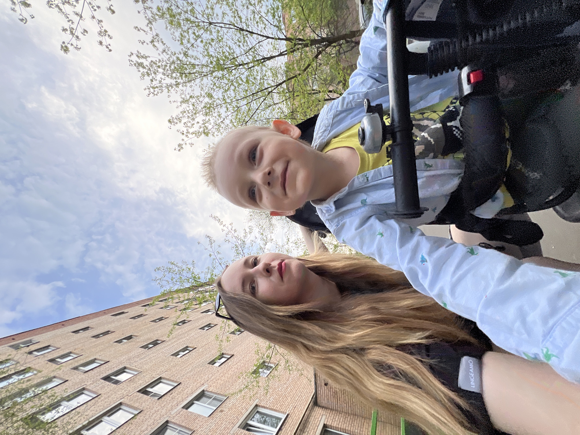 A woman and a young boy are visible in the image, with a tall brick building and sky in the background. The boy is in a wheelchair or stroller, indicated by the visible frame and attachments. He has blonde hair and is wearing a yellow shirt under a light blue collared shirt. The woman, likely the boy's mother, has long blonde hair and is wearing sunglasses on top of her head. The building is a multi-story brick structure with rows of windows. The sky is partly cloudy, with blue patches visible. The presence of new leaves on nearby trees suggests the season is spring. The location is the city of Mytishchi, Russia.