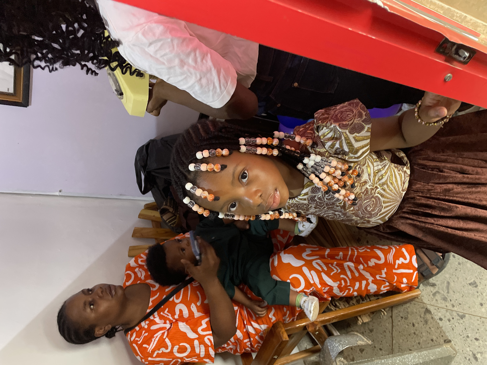 The image depicts several individuals, likely family members, in an indoor setting. A woman is seated, wearing an orange and white patterned top, holding a young child dressed in green. A young girl with beaded braids stands nearby, wearing a patterned blouse and a brown skirt. Another individual is partially visible in the background, holding a yellow object, possibly a telephone. The location is identified as Barakin Akawo, Nigeria. Other details include the presence of seating, clothing patterns, and various objects within the environment.