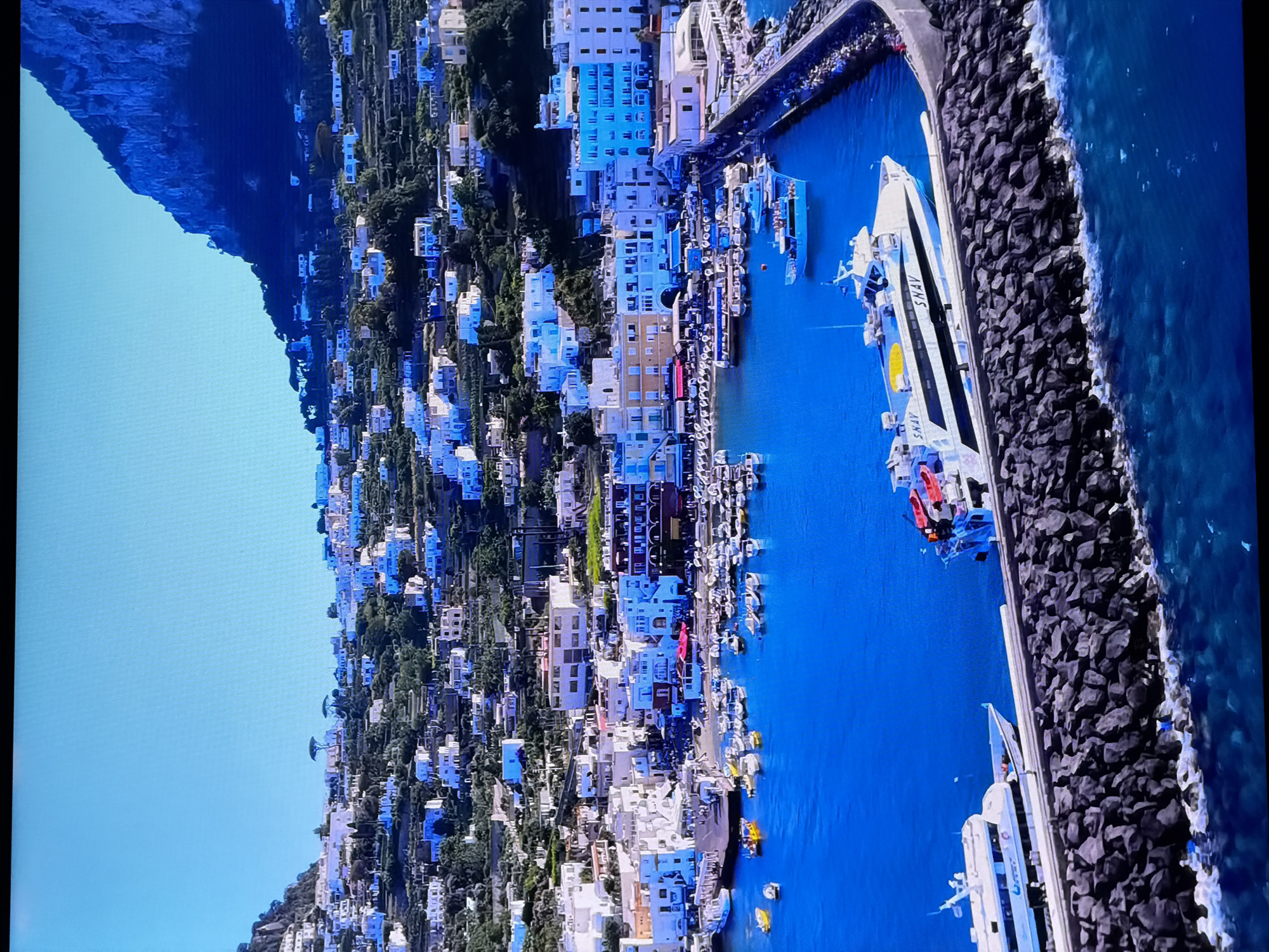 This image depicts a scenic harbor filled with numerous boats and ferries, situated at the base of a steep, terraced hillside covered with white buildings. The water is a vibrant blue, and the sky is clear and bright. Several white buildings with blue accents line the waterfront, with more structures cascading up the hillside. A large ferry with "SNAV" written on its side is docked at the pier, along with other smaller vessels. The hillside is densely populated with houses and vegetation, creating a picturesque coastal town. There are no visible people in the image. The location described is the city of Chirchik, Uzbekistan.