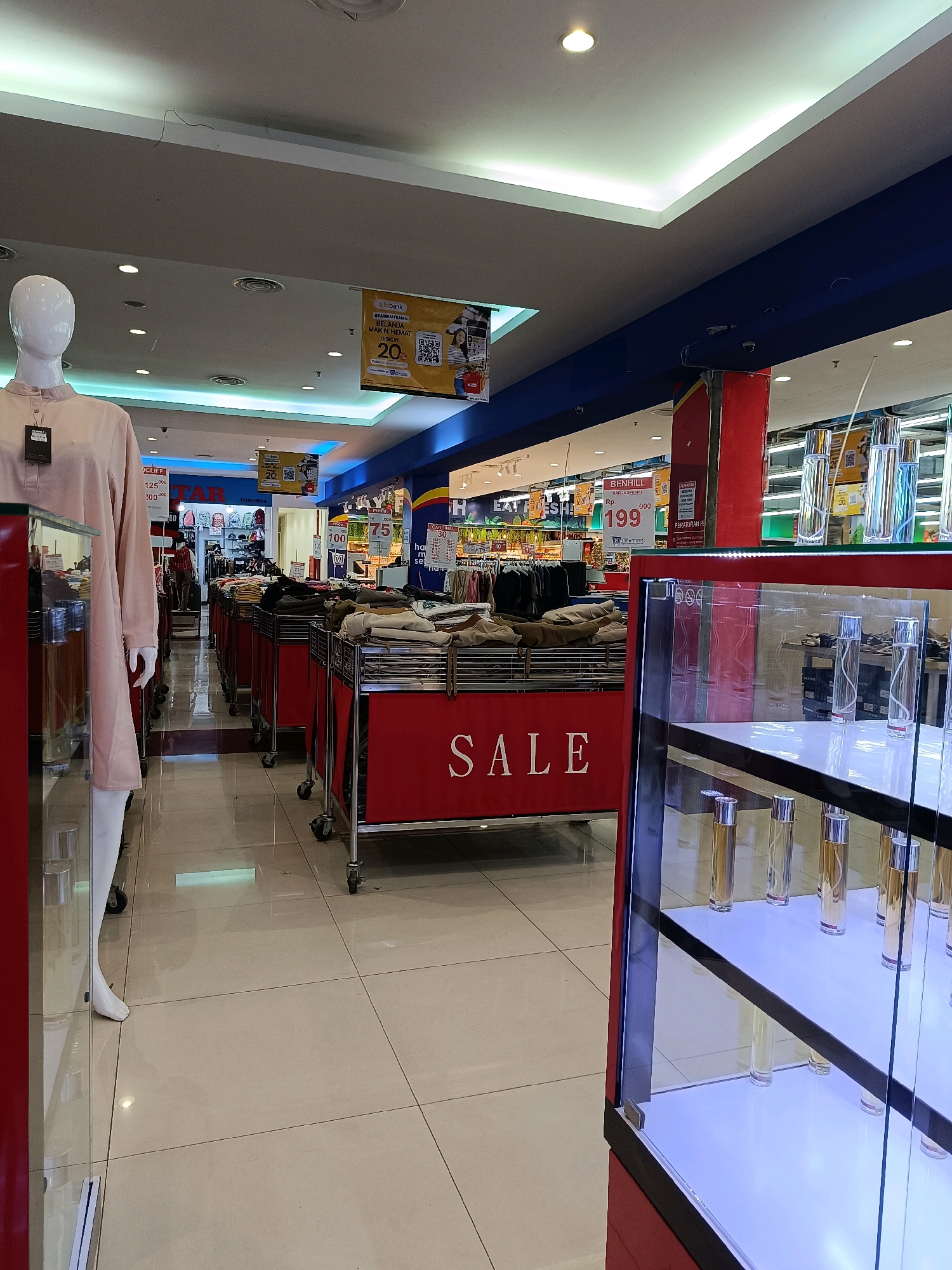 The image depicts an interior view of a retail store, possibly a department store or supermarket, in Tomohon, Indonesia. The store features a well-lit environment with a tiled floor and a suspended ceiling. In the foreground, a mannequin dressed in a light pink garment is displayed. To the right, a glass display case showcases rows of perfume bottles, illuminated from below.
In the midground, rows of red display racks are filled with folded clothing items. A prominent red banner on one of these racks reads "SALE" in white capital letters. Beyond the clothing racks, more merchandise is visible, including what appears to be clothing and other retail items. Several signs are posted throughout the store, some with promotional offers and prices. One sign, partially visible, indicates "BENHILL" and a price of "199". Another sign reads "CARDINAL" with a discount of "50%". Further back, aisles of products are organized, suggesting a larger retail space.
The overall scene suggests a commercial environment focused on clothing and cosmetic sales, with current promotions being highlighted. The lighting and store layout are typical of a modern retail establishment.