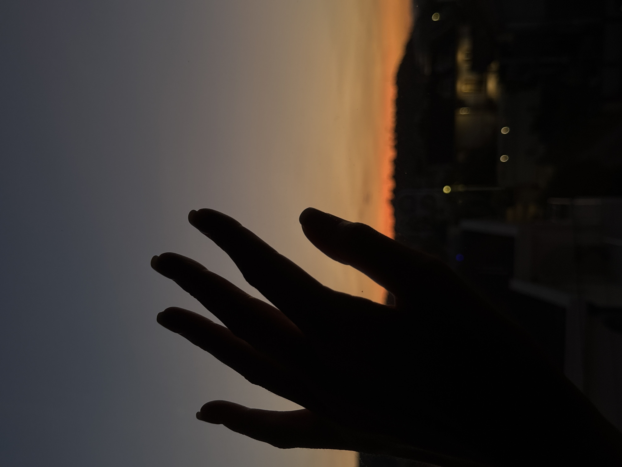A silhouette of a hand is in the foreground. The background shows a gradient sky with hues of gray, orange, and dark red. In the upper-right corner, a darkened area contains blurred light sources, likely suggesting city lights. While it's difficult to pinpoint the location with absolute certainty, the scene suggests an elevated perspective, possibly from a building in Podgorica, Montenegro, capturing a sunset or sunrise view.