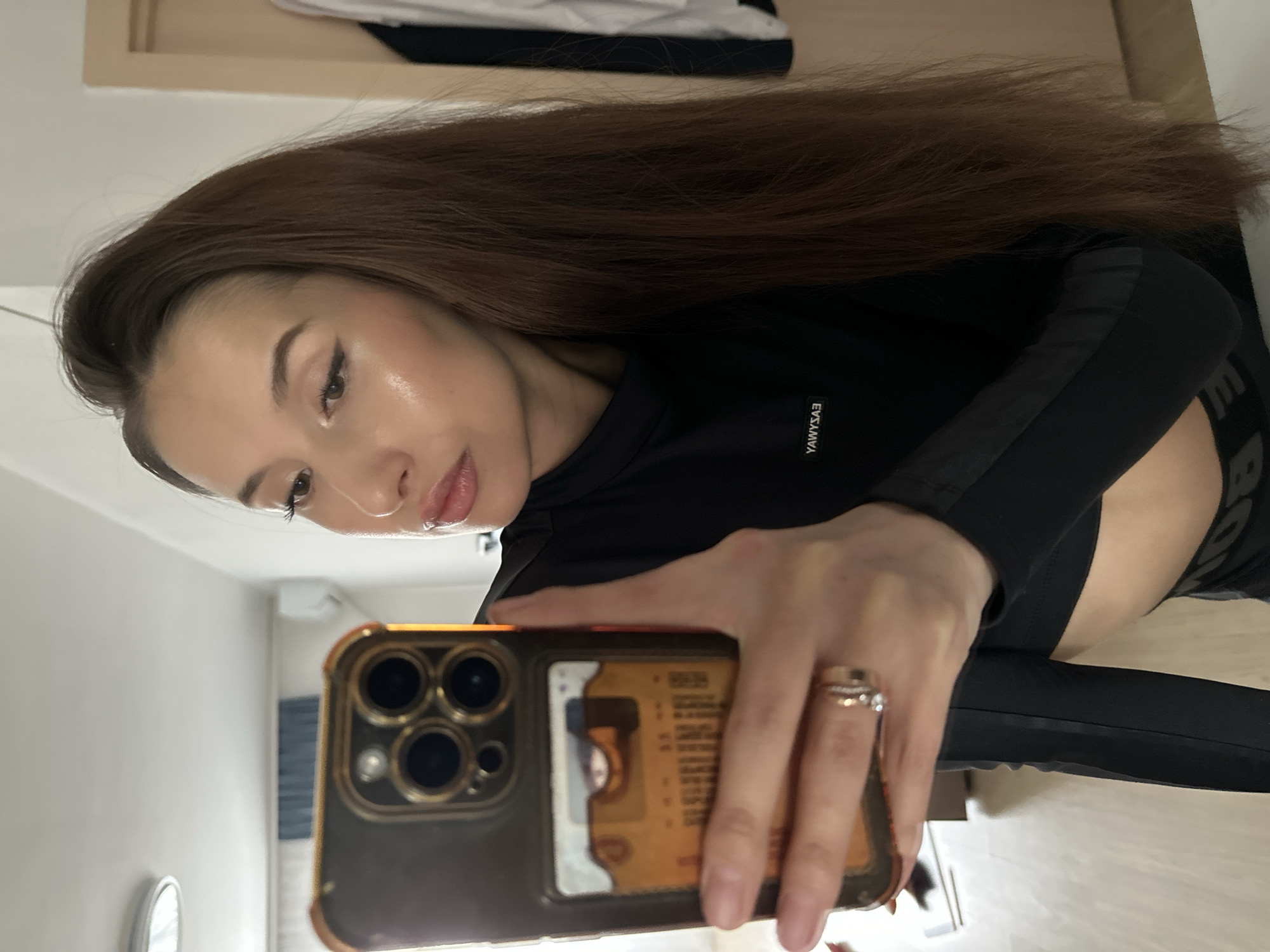 A woman is taking a selfie indoors. She is wearing a black long-sleeved top. Her hair is long and brown. She is holding a smartphone with a gold case in her right hand. A ring is on her finger. The background includes a glimpse of a room, possibly a bedroom or living space. It is not possible to determine the location as Yekaterinburg, Russia from the information provided in the image.