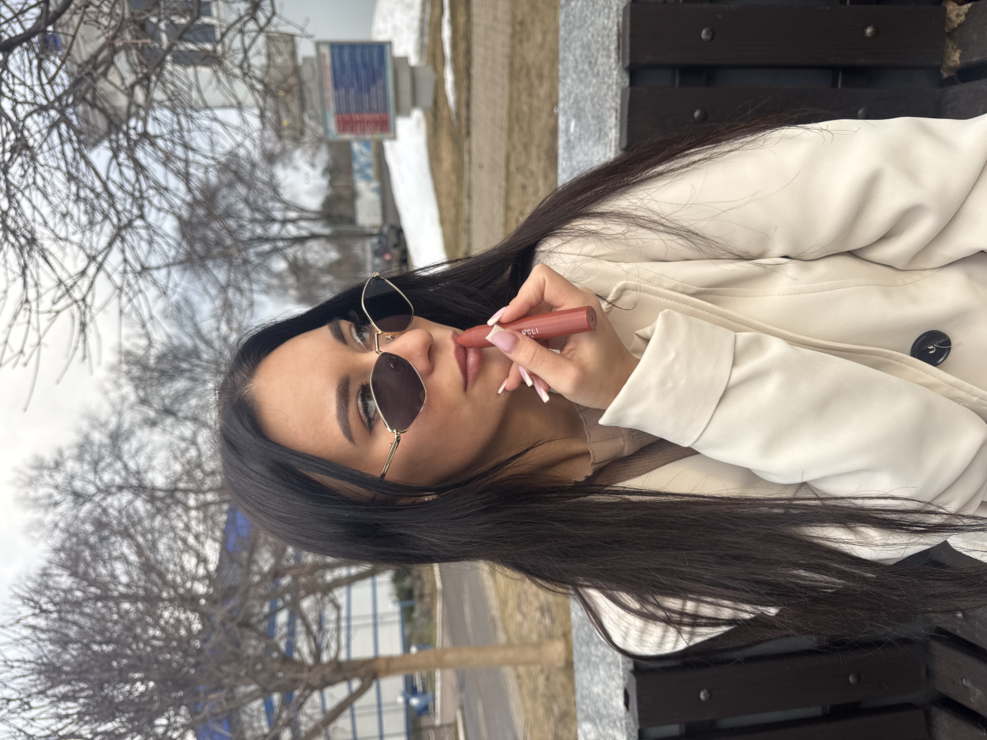 A woman with dark hair and sunglasses is applying lipstick. She wears a white jacket and sits on a dark bench. Outside, there are trees, and in the background, a building. The location is Chaikovsky, Russia.