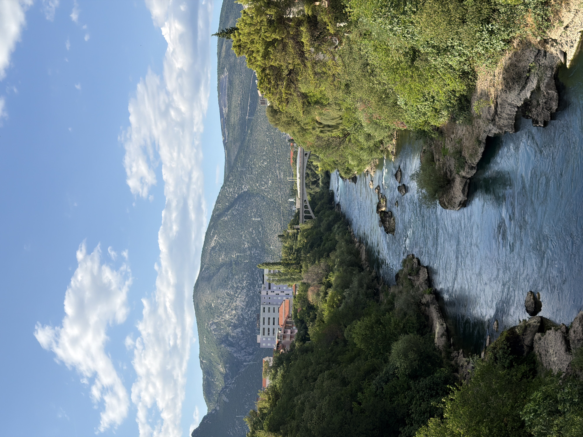 The image depicts the Neretva River flowing through the city of Mostar, Bosnia and Herzegovina, with lush green hills forming the backdrop. The river is characterized by its clear blue water and visible rocks scattered throughout its course. On the left bank, a modern multi-story building with a grey facade and several windows is situated amidst trees. Adjacent to it are smaller buildings with terracotta roofs. Further upstream, a white arched bridge spans the river. The right bank is densely covered with various types of green trees and foliage. The sky above is a vibrant blue, adorned with fluffy white clouds. The scene is captured from an elevated perspective, offering a panoramic view of the natural landscape integrated with urban elements.