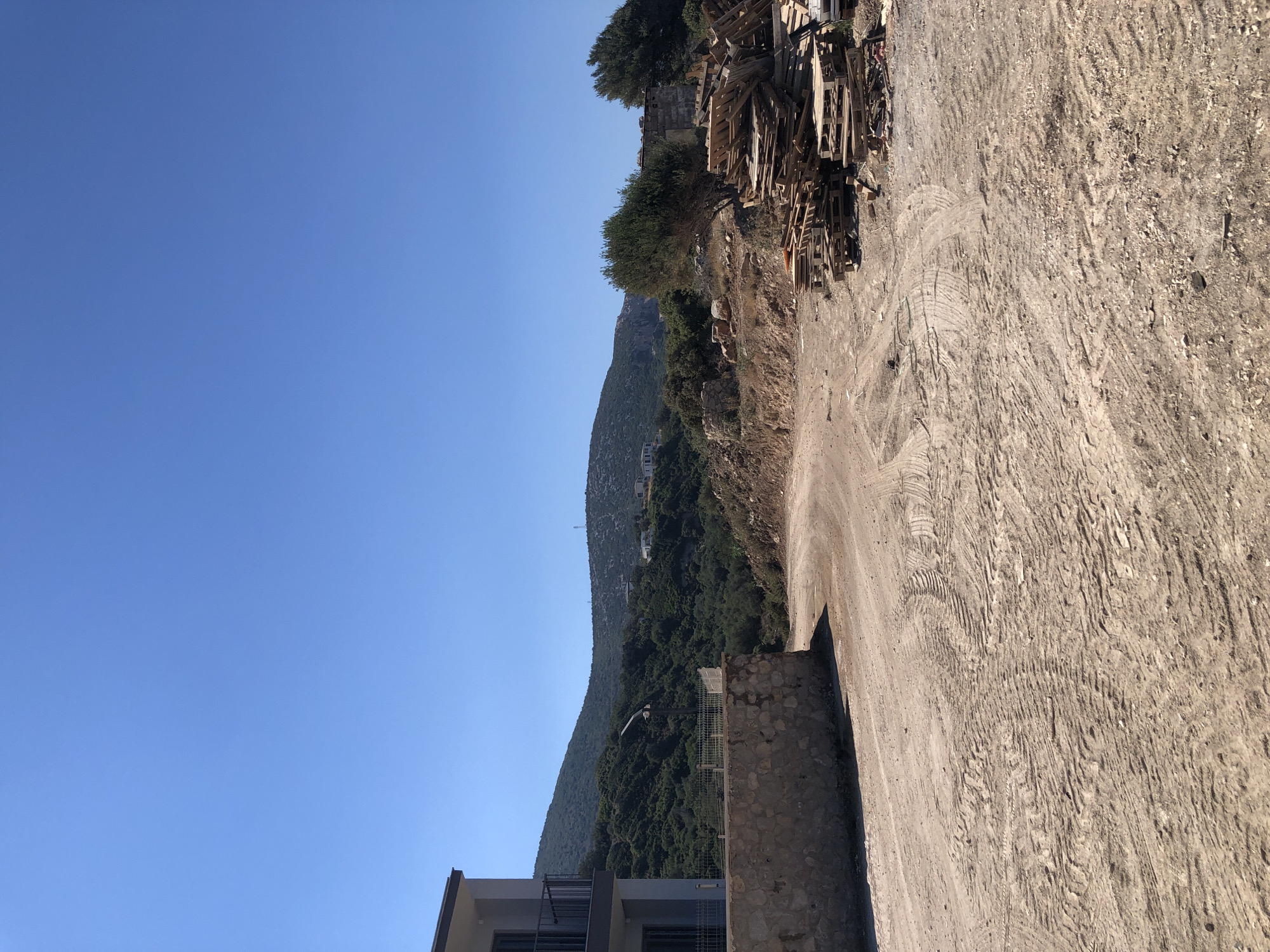The image depicts an outdoor scene in Karaburun, Turkey, under a clear blue sky. In the foreground, a dirt road is visible, marked with tire tracks from vehicles. To the left, a modern building with a balcony and dark railings stands beside a low stone wall. Behind the wall, a fenced area is apparent. Further into the scene, a pile of wooden pallets is stacked on the right side of the dirt road. The background features a lush, green hillside dotted with scattered buildings, and a distant mountain range under the bright sky. The overall impression is of a construction or development area in a semi-rural or coastal environment.