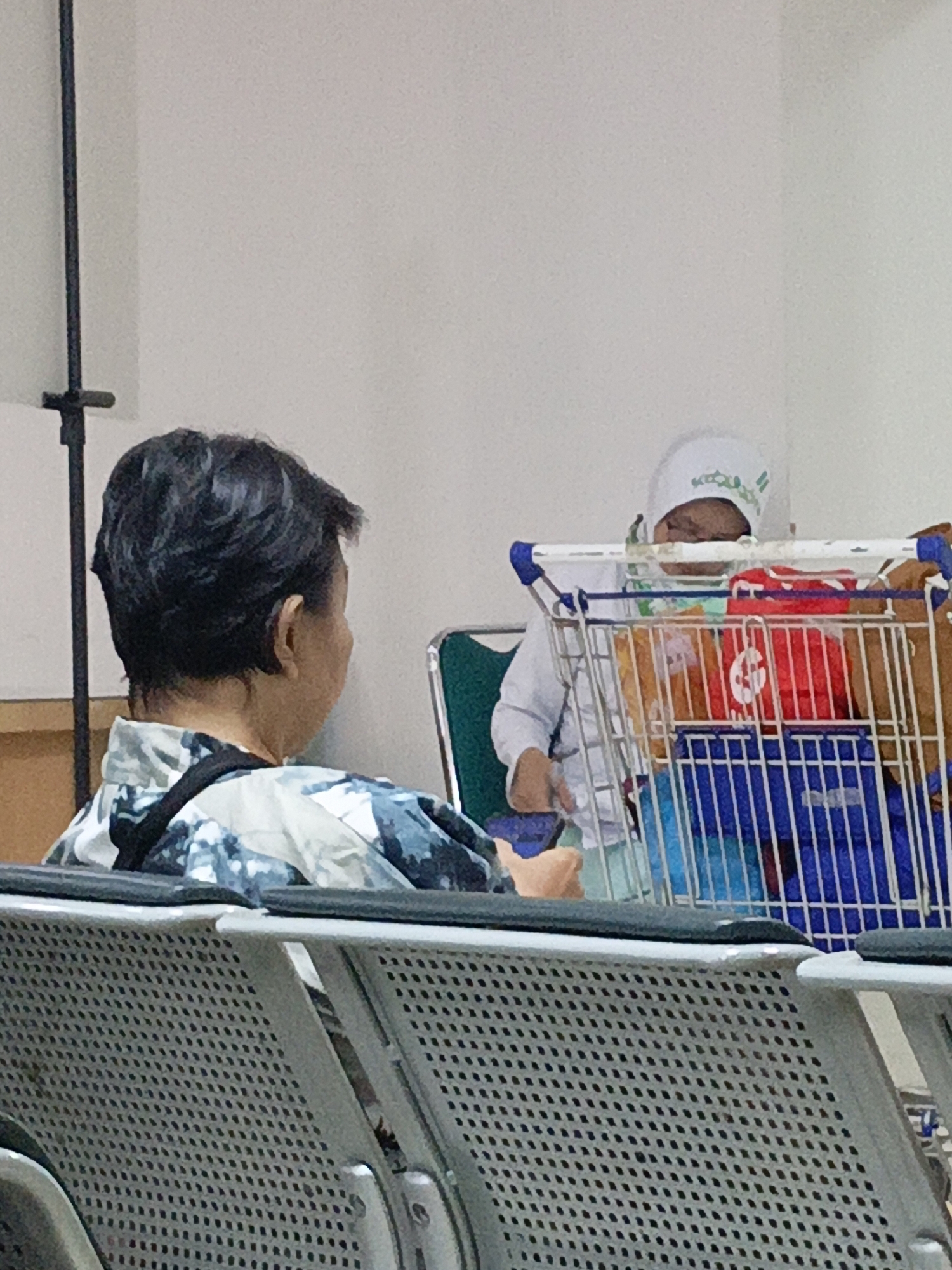 An adult with short, dark, wavy hair wearing a patterned shirt is seated facing away from the camera. In the background, a young child wearing a white cap and white clothing is sitting in a shopping cart. The child's face is partially obscured. The shopping cart contains various items, including red and brown bags. The scene appears to be an indoor waiting area with rows of gray seating in the foreground. The overall lighting is neutral, suggesting an indoor environment in East Jakarta, Indonesia.