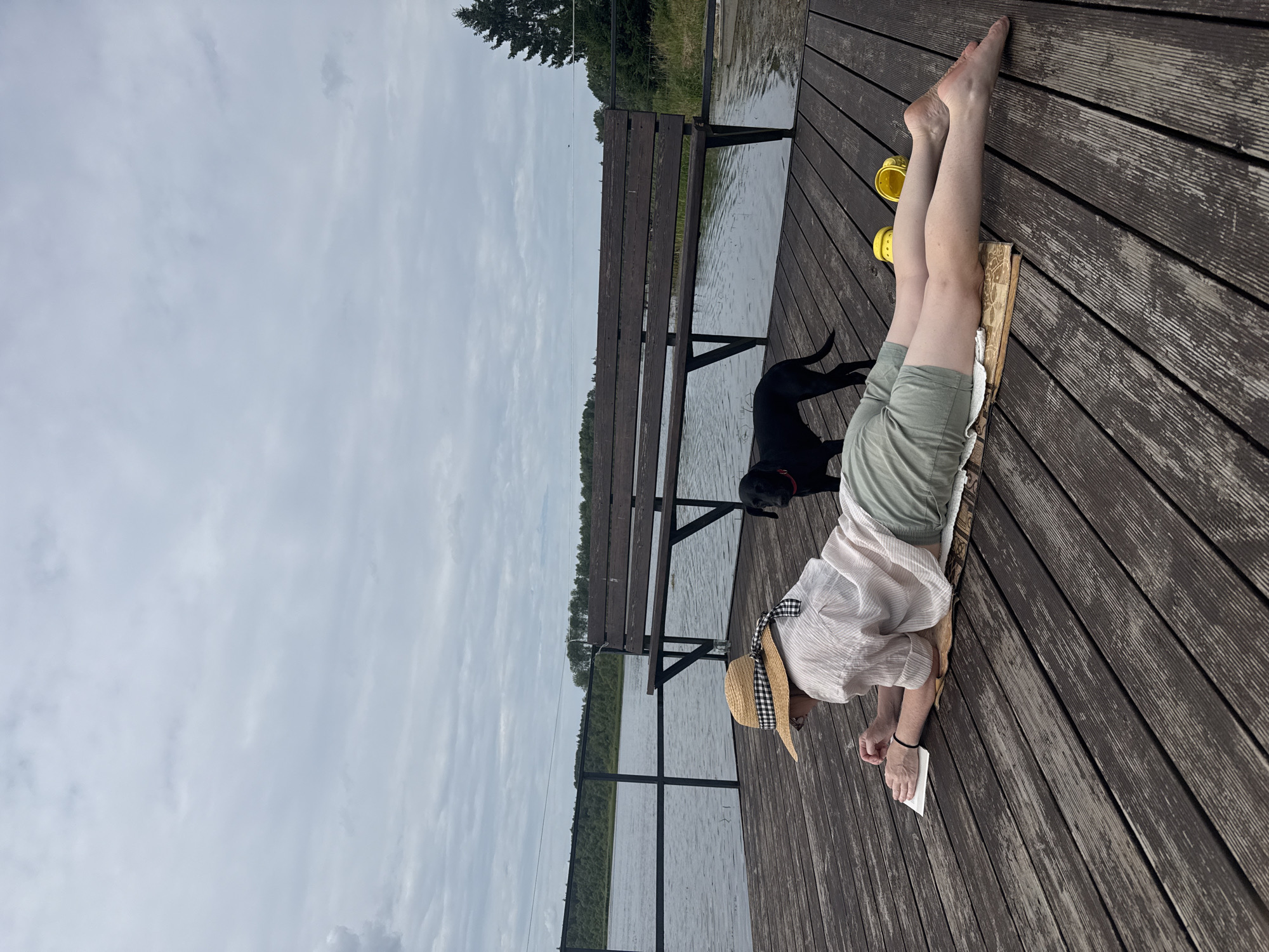 An outdoor, overcast, medium shot captures a person lying prone on a wooden dock by a body of water. The person, facing away from the camera and towards the left, wears a straw hat with a checkered band, a light-colored, short-sleeved shirt, and olive-green shorts. They are holding a small, open book and appear to be reading. Next to the person's feet are two yellow clogs. A black dog stands behind the person, facing left. The wooden dock, constructed from planks, extends into the water. A dark wooden bench is positioned on the dock behind the person and dog. A railing, with vertical posts and horizontal bars, runs along the edge of the dock near the water. The water is rippled and appears calm, reflecting the overcast sky. A treeline borders the far side of the water, and a tall evergreen tree is visible on the right side of the frame. The location is the city of Chikino, Russia.