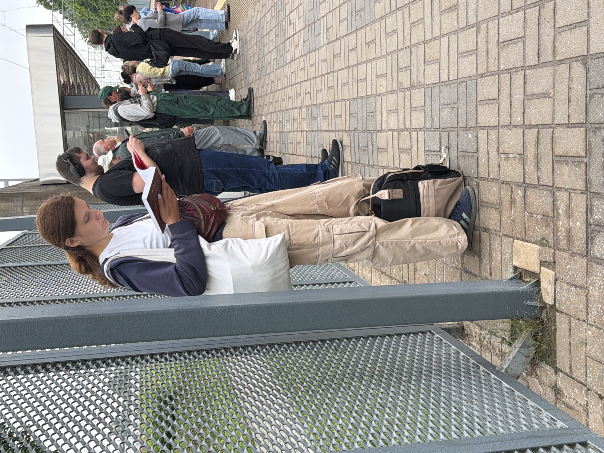 The image depicts a group of people standing on a paved platform, likely a train station or bus stop, in Moscow, Russia. The ground is made of rectangular stone pavers arranged in a herringbone pattern. A metal fence with a diamond-shaped mesh pattern runs along the left side of the frame, supported by a gray metal pillar. The people are standing in a line, facing towards the right. The primary subject, a woman in the foreground, is wearing tan cargo pants and a dark blue jacket over a white t-shirt. She is holding an open book and appears to be reading. A white canvas bag is slung over her shoulder, and a dark backpack with tan accents rests at her feet. Other individuals in the background are also engaged in various activities, such as looking at their phones or reading. The overall atmosphere suggests a public waiting area, possibly during an overcast day.