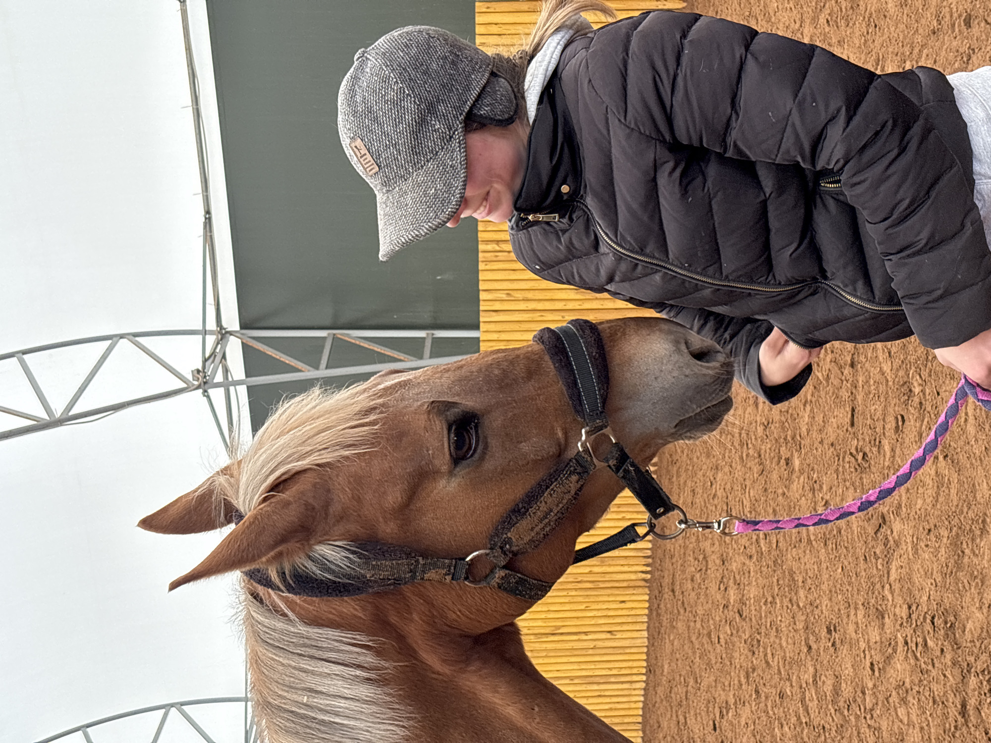 A fair-skinned woman wearing a black puffer jacket and a gray tweed cap is interacting with a horse. The horse is light brown with a white mane and is wearing a black halter. The woman is holding a pink and purple braided lead rope attached to the halter. The background consists of a metal structure, a green tarp or wall, and wooden slats. The ground is brown. The location is Русский Вожой, Russia.