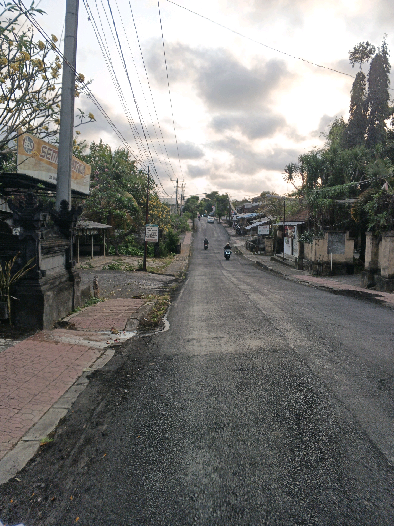 The image depicts a street scene in Sukawati, Indonesia, on an overcast day. The perspective is from ground level, looking up a paved road that slopes slightly uphill.
On the left side of the road, there is a sidewalk made of red pavers, bordered by a grey concrete curb. Adjacent to the sidewalk is a dark, ornate stone wall and gate, typical of Balinese architecture. Behind the wall, there are buildings with corrugated metal roofs, some with open-air structures. A tall utility pole with numerous wires runs along the left side of the road, near the ornate gate. A large sign with the text "SEMA" and other writing is visible on a structure to the left. Yellow flowers bloom on trees and bushes throughout the scene.
The road itself is asphalt and appears to be in moderate condition, with some patches of darker, wet-looking asphalt and scattered leaves. Several motorcycles are visible on the road. Two motorcycles are in the foreground, one with a rider, and another a short distance ahead. Further up the road, another motorcycle and a car are visible in the distance.
On the right side of the road, there are more buildings with similar tiled or corrugated roofs. Some structures have walls constructed of stone or concrete blocks. More utility poles and wires are present on this side of the road, further up the incline. Lush green vegetation and trees are abundant on both sides of the street, contributing to a tropical atmosphere. The sky is filled with heavy, grey clouds, with a bright patch of light suggesting the sun is attempting to break through. The overall impression is a moderately busy street in a residential or commercial area of Sukawati, with a blend of traditional and modern elements.