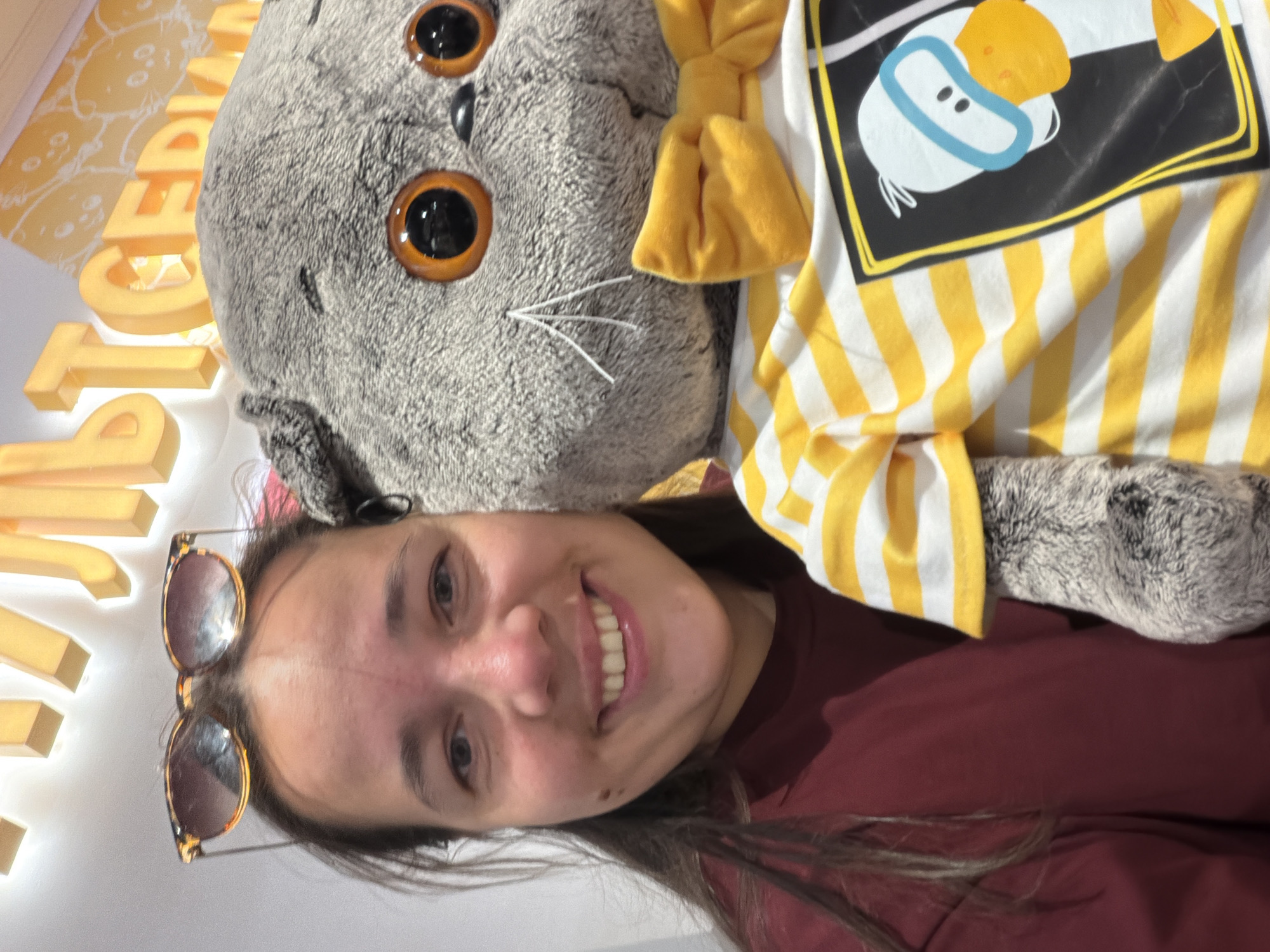 The image shows a selfie taken by a woman with a large, gray, stuffed cat wearing a yellow bow tie and a striped yellow and white shirt. The shirt features a cartoon duck graphic. The woman has dark hair and is wearing sunglasses perched on her head and a maroon shirt. The background includes partially visible yellow lettering on a white wall. The setting appears to be a store or promotional area, possibly located in Moscow, Russia, given the context and potential brand affiliations associated with the plush toy. The primary interaction is the woman taking a selfie with the stuffed cat.