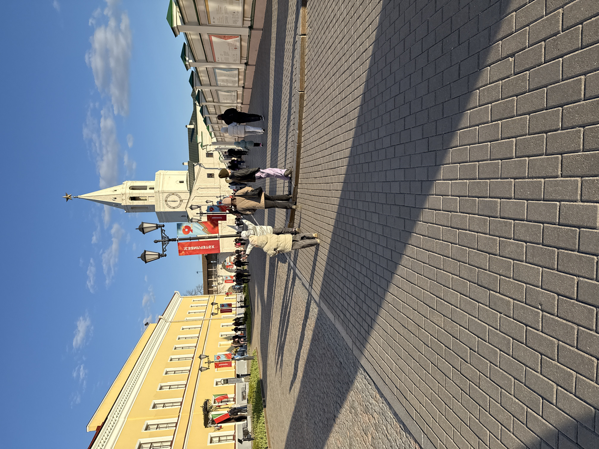 The image depicts a street scene in Kazan, Russia, under a clear, blue sky with scattered clouds. Prominently featured is the Soyembika Tower, a distinctive, leaning architectural landmark of the Kazan Kremlin, casting a shadow on the cobblestone pavement. Pedestrians are strolling along the brick paved walkway. Adjacent to the walkway is a yellow building. Along the walkway are street lamps with banners attached to them.