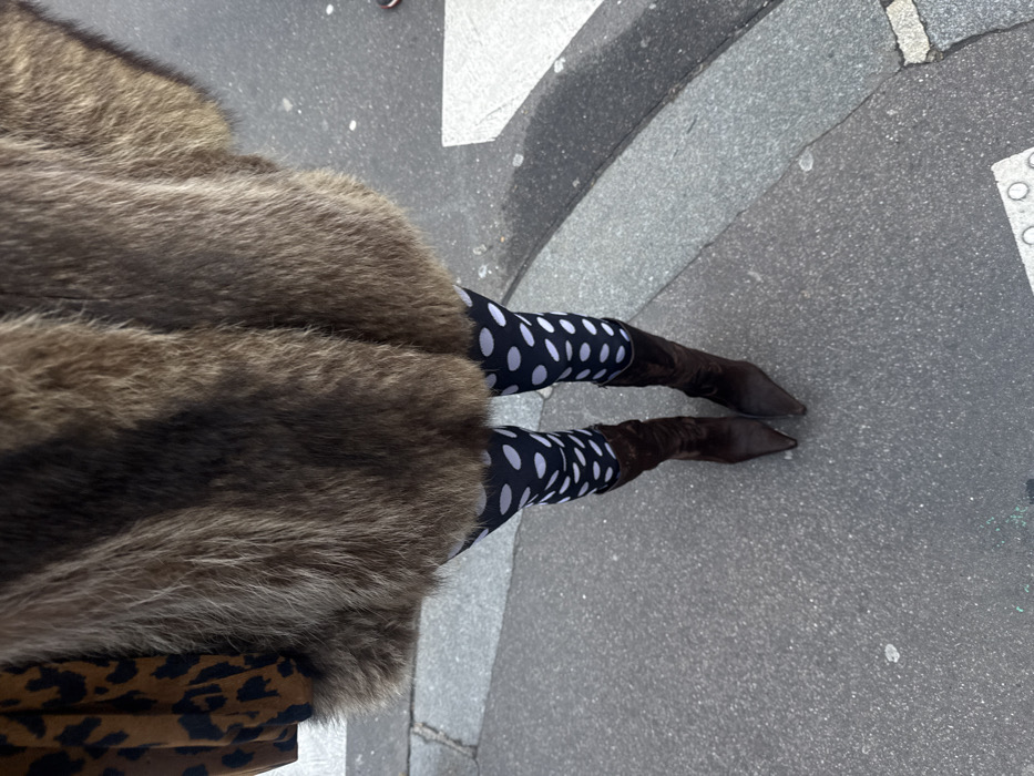 From a high angle, a figure clad in a large, brown fur coat and black leggings with white polka dots stands on a grey pavement. The leggings are tucked into knee-high, brown suede boots with pointed toes. A hint of a brown and black leopard print garment is visible beneath the fur coat on the left. The surrounding pavement shows some white markings and a curved edge.