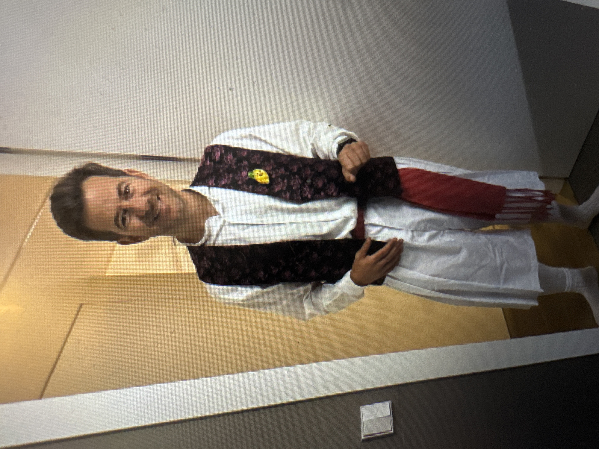 A man in traditional Spanish attire stands in what appears to be a hallway, with a door on the left and a wall on the right. He is wearing a white, long-sleeved shirt, a dark patterned vest, white wide-leg pants, and white socks. A red sash with tassels is wrapped around his waist. He has dark hair and is smiling at the camera. The environment suggests an indoor setting, possibly a residence or a public building. The image quality is grainy, suggesting it was taken with a phone camera or is a screenshot. There are no explicit indicators of a specific location within Murcia, Spain.