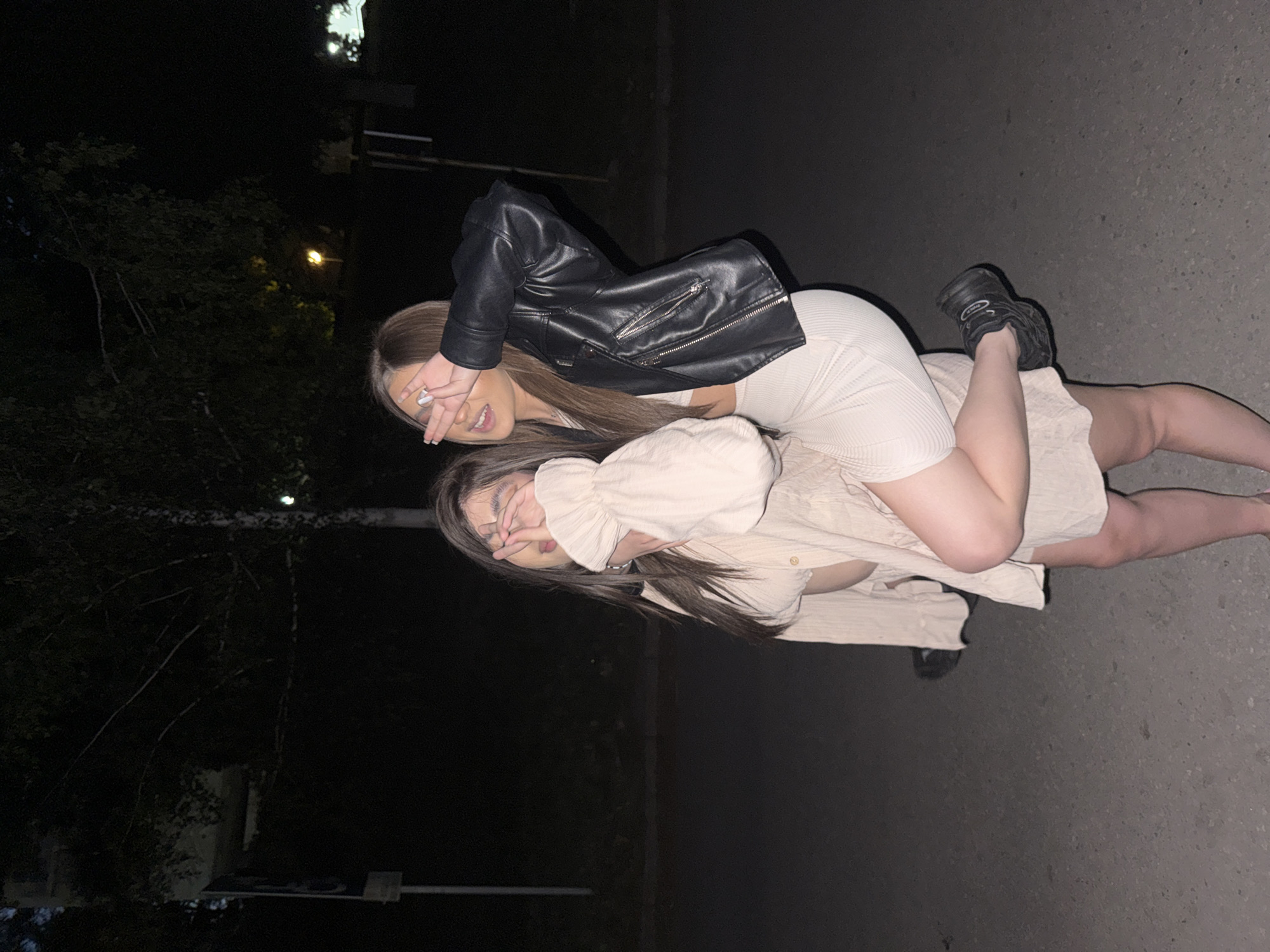 This image captures two young adult females posing outdoors at night in an urban area of Blagoevgrad, Bulgaria. The specific location within the city is not discernible due to a lack of identifiable landmarks.
The subject on the left, with dark, long hair, is wearing a light beige textured button-down shirt with ruffled cuffs, unbuttoned to reveal a lighter top underneath. Her lower attire is partially obscured, but she appears to be wearing light-colored open-toed footwear. Her left hand is raised, covering her eyes with fingers splayed in a "V" shape, while her mouth is open, indicative of a smile or laugh. She supports the second subject, with her right arm around the other's waist or leg.
The subject on the right, with long, brown hair with lighter highlights, is wearing a black leather motorcycle-style jacket over a light-colored, ribbed crop top and a matching ribbed, fitted mini-skirt. She is wearing black chunky sneakers. Her right hand is raised, covering her eyes in a "V" shape, mirroring the first subject's pose. Her left leg is bent at the knee and raised, suggesting she is either being held or playfully perched on the first subject, with her left arm around the first subject's shoulders. Both subjects exhibit expressions consistent with smiling or laughing.
The environment is dark, indicating nighttime conditions. The ground surface is paved, likely asphalt or concrete. In the background, silhouetted trees with sparse foliage are visible against the dark sky. Multiple artificial light sources, including a distinct warm yellow streetlight, are present in the mid-ground and background, casting ambient light on the scene. A vertical pole, consistent with a signpost, is visible in the left background.