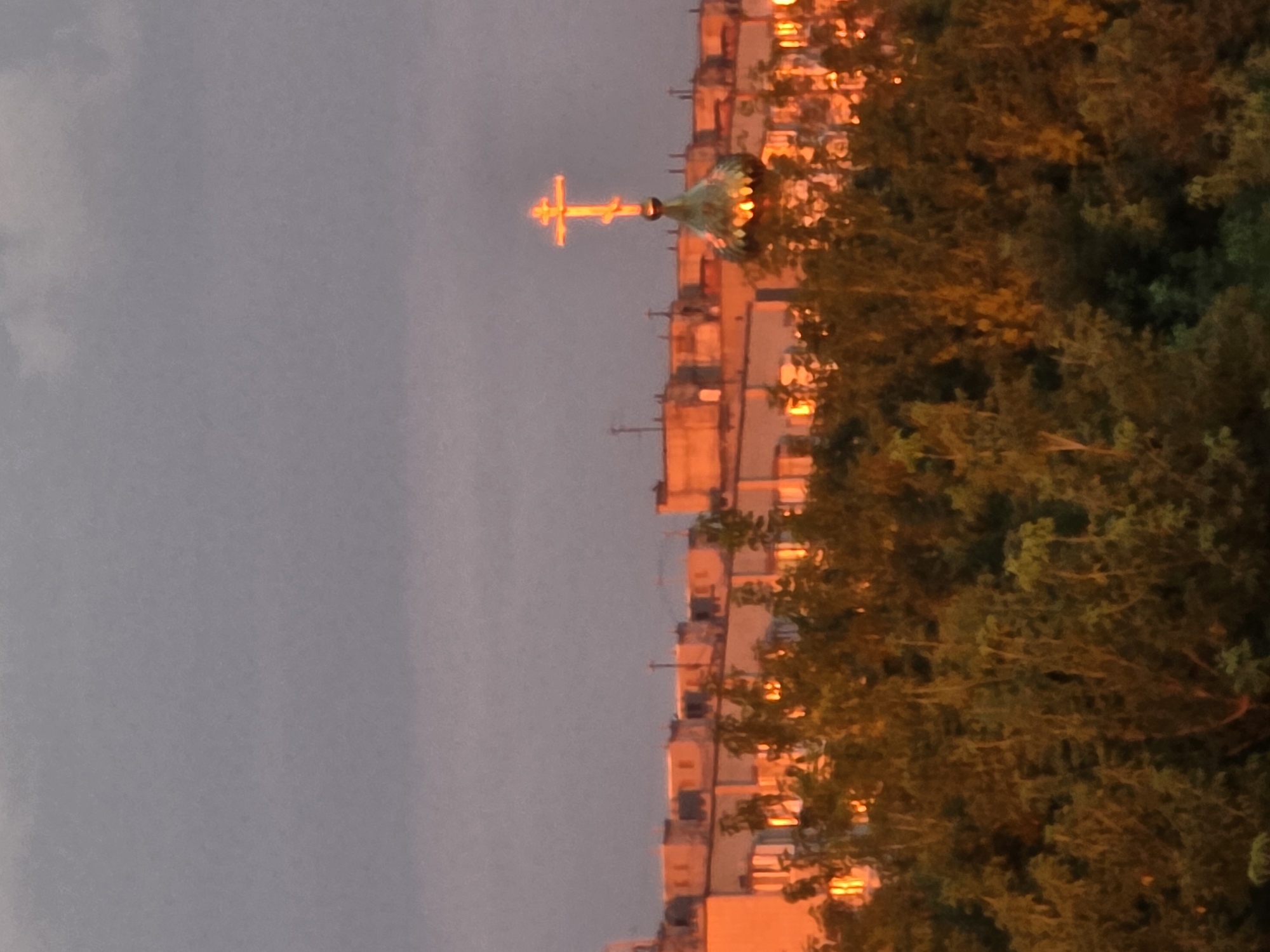 The image depicts an urban landscape at dusk, possibly in Chaikovsky, Russia, characterized by an overcast sky tinged with pink and orange hues from the setting sun. In the mid-ground, a line of apartment buildings is visible, their facades illuminated by the warm light. Several windows reflect the sunset, creating a glowing effect. Above the buildings, the golden dome of an Orthodox church topped with a bright, illuminated cross stands out against the dusky sky. In the foreground, a dense canopy of trees, with foliage reflecting the sunset's colors, partially obscures the view of the buildings and the church. The overall atmosphere is serene and tinged with the warm, diffused light of twilight.