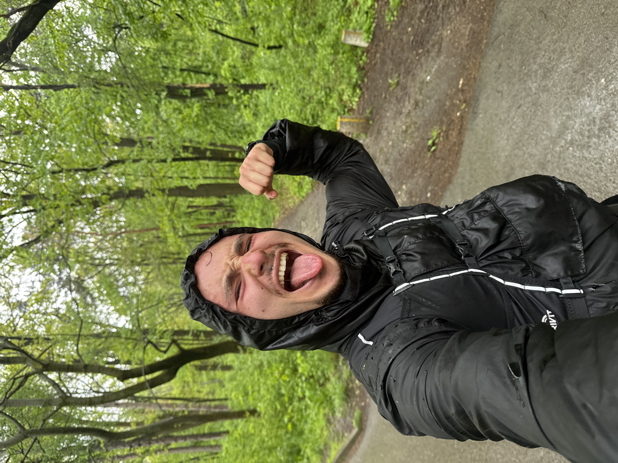A playful selfie captures a man outdoors in a park or forest in Sofia, Bulgaria, likely during or after rainfall. The subject, a male with short dark hair and a beard, wears a black hooded, water-resistant jacket with white stripe details. He exhibits an energetic and exaggerated expression, with his mouth wide open, tongue sticking out, and eyes scrunched up, while flexing his right bicep. The background features dense, lush green foliage and a wet path, indicating damp conditions from recent rain. Water droplets are visible on his jacket. Partially visible, mirrored text resembling "RIVIT" can be seen on the lower left side of his jacket. The image conveys a lighthearted and resilient mood, embracing the outdoor experience despite the rainy weather.
