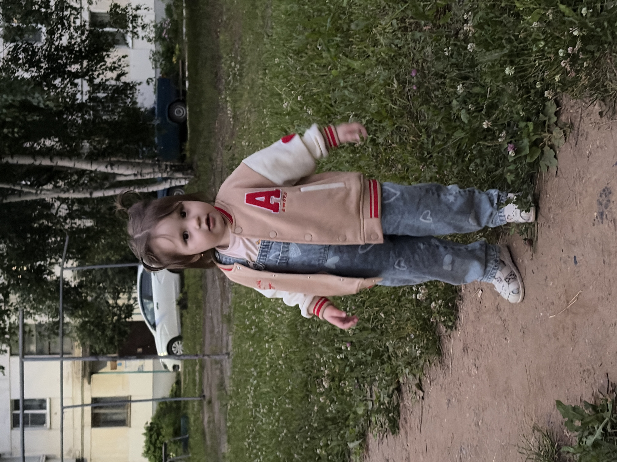 A young girl stands on a dirt path in a grassy area outside a residential building. The girl, with brown hair tied up, is wearing a light pink shirt, denim overalls with heart patterns, and a tan and cream colored baseball jacket with red trim and an "A" applique on the left chest. She also wears white sandals. In the background, a white car is parked near a light-colored apartment building with visible windows. To the right of the building, trees and another parked car, which is dark blue, are visible. The ground is covered in green grass with small white flowers.