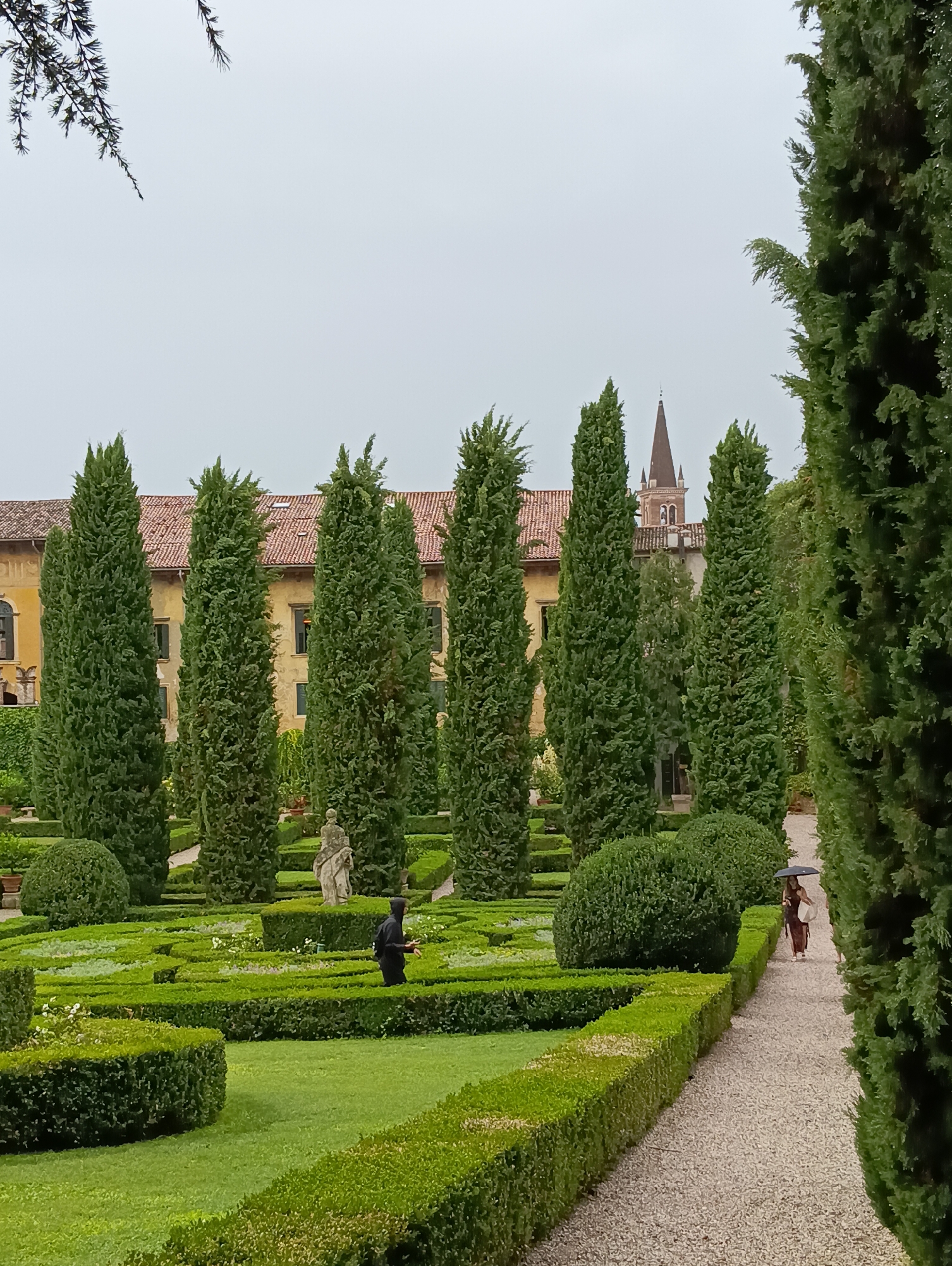 The image depicts a manicured garden with geometric hedges and tall cypress trees, likely located in Verona, Italy. In the background, a building with a terracotta roof and arched windows is visible, along with a church spire. A stone statue stands amidst the greenery. Two people are present in the scene: one individual in a black hooded jacket walks through the garden, and another person, possibly a woman in a dark dress, walks on a gravel path holding a black umbrella. The sky is overcast, suggesting a cloudy day. The overall impression is one of a well-maintained historical garden.