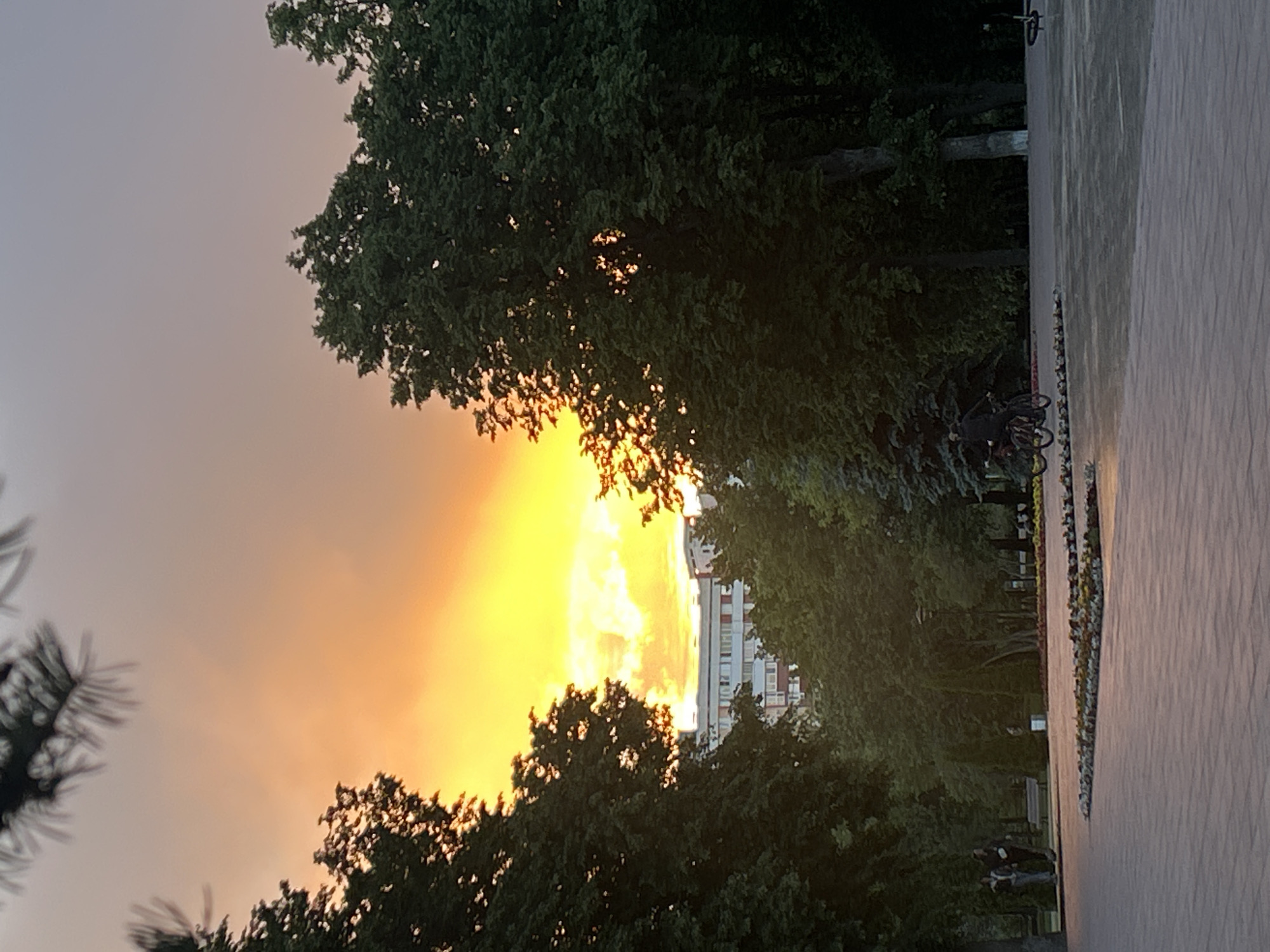 The image shows a sunset in Tolyatti, Russia. The sun is low on the horizon, creating a bright yellow and orange glow in the sky that peaks between the silhouettes of trees. A building can be seen in the distance, framed by the trees and sunset. On the right is a paved walkway with planted flower beds along the edge and people walking along it. Several bicycles are parked next to the walkway.