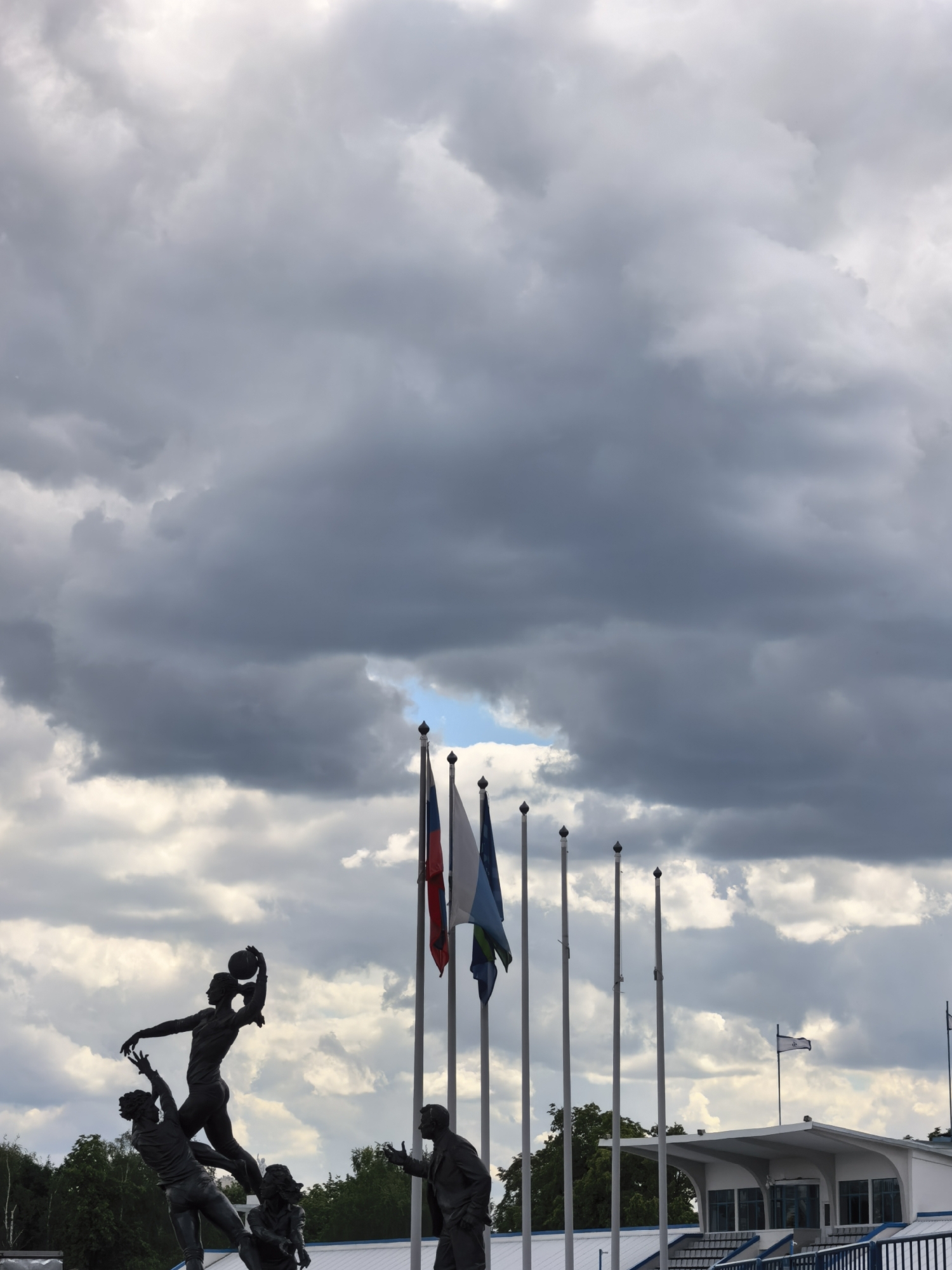 A statue depicting basketball players is erected in front of several flagpoles, with a building in the background, under a cloudy sky in Unknown, Unknown. The statue features a female basketball player reaching for a ball held above her head, while two male players appear to be reaching for her. Another figure stands to the right, seemingly observing. Several flagpoles stand behind the statue, with flags of varying colors and designs. The building in the background is white with blue accents and has a modern architectural style. The sky is overcast with heavy, dark clouds.