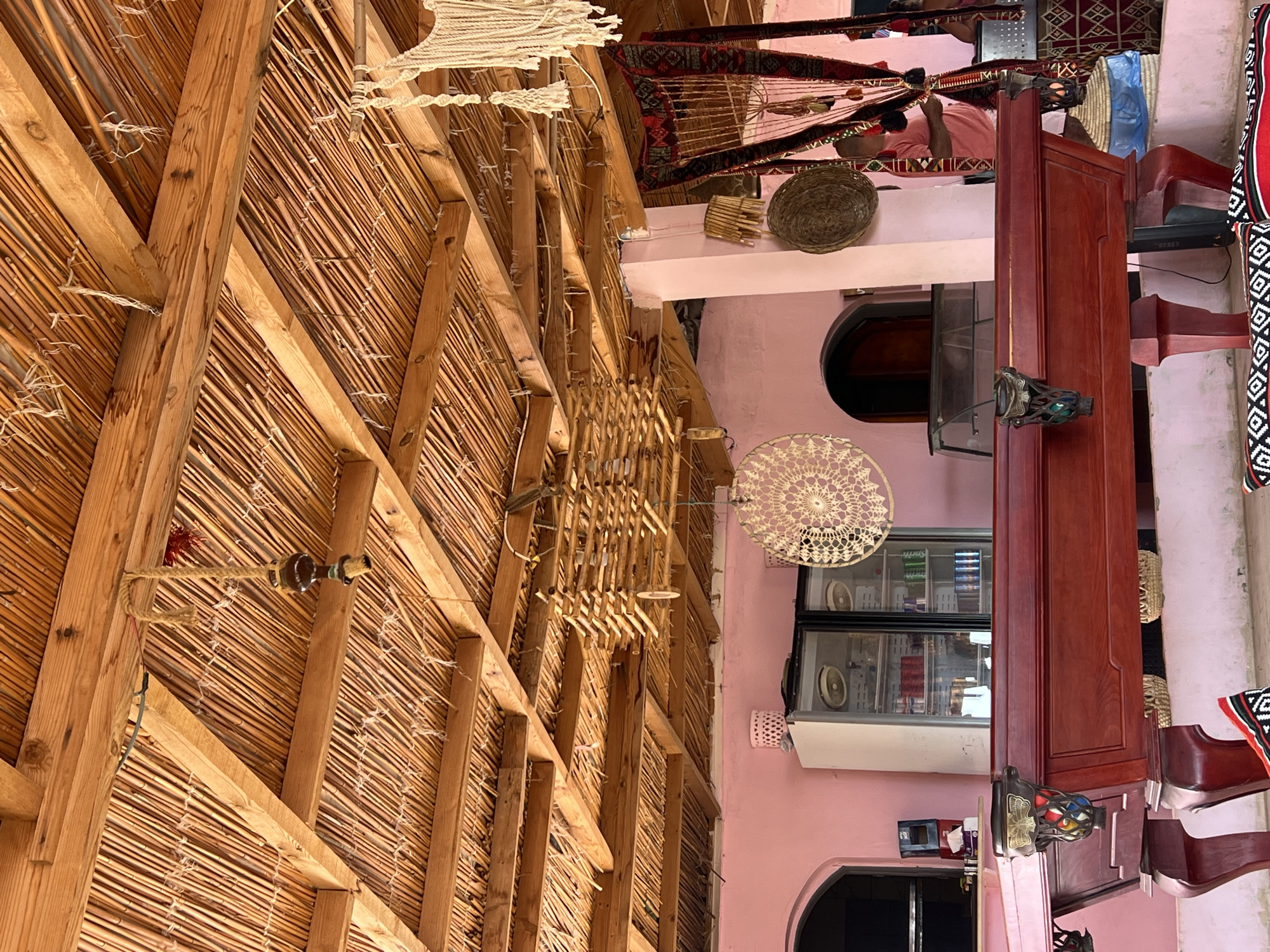 The image depicts an indoor scene, likely a cafe or lounge, with a rustic and bohemian aesthetic. The ceiling is constructed of natural reeds and wooden beams, creating a warm, textured canopy. Hanging from the ceiling are decorative elements including a macrame wall hanging, a wooden mobile, and a hanging lamp with a ceramic shade. The walls are painted a vibrant pink.
In the foreground, a dark red wooden pool table with ornate metal pockets holding colorful billiard balls is prominently featured. The table has intricately carved legs and detailing. In the background, a large glass-doored refrigerator filled with beverages is visible. To the right of the refrigerator, a man wearing a pink t-shirt is partially visible, standing near a tapestry or rug hanging on the wall. Additional decorative items, such as woven baskets and circular woven wall hangings, are scattered throughout the space. The floor appears to be concrete, with patterned cushions placed in front of the pool table. The overall impression is of a casual, eclectic, and inviting recreational space. The image does not contain any explicit indicators of its specific location within Egypt or any city.