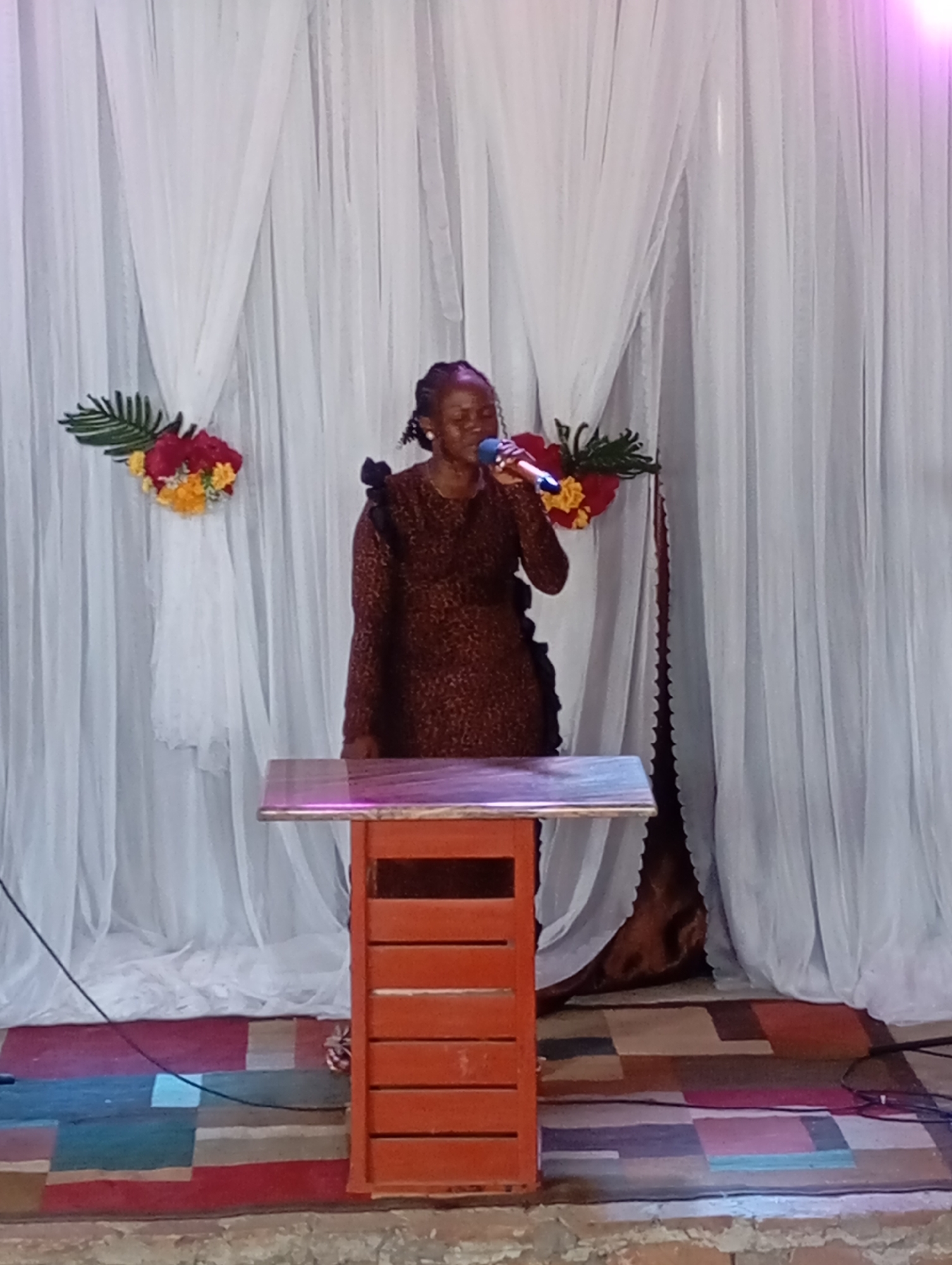 A woman stands at a wooden podium, holding a microphone and speaking. She has her hair in braids and is wearing a brown patterned dress. Behind her is a backdrop of white draped fabric adorned with floral arrangements on either side. The podium is orange with a light-colored top. The floor is covered with a colorful patchwork rug. The location is Uke, Nigeria.