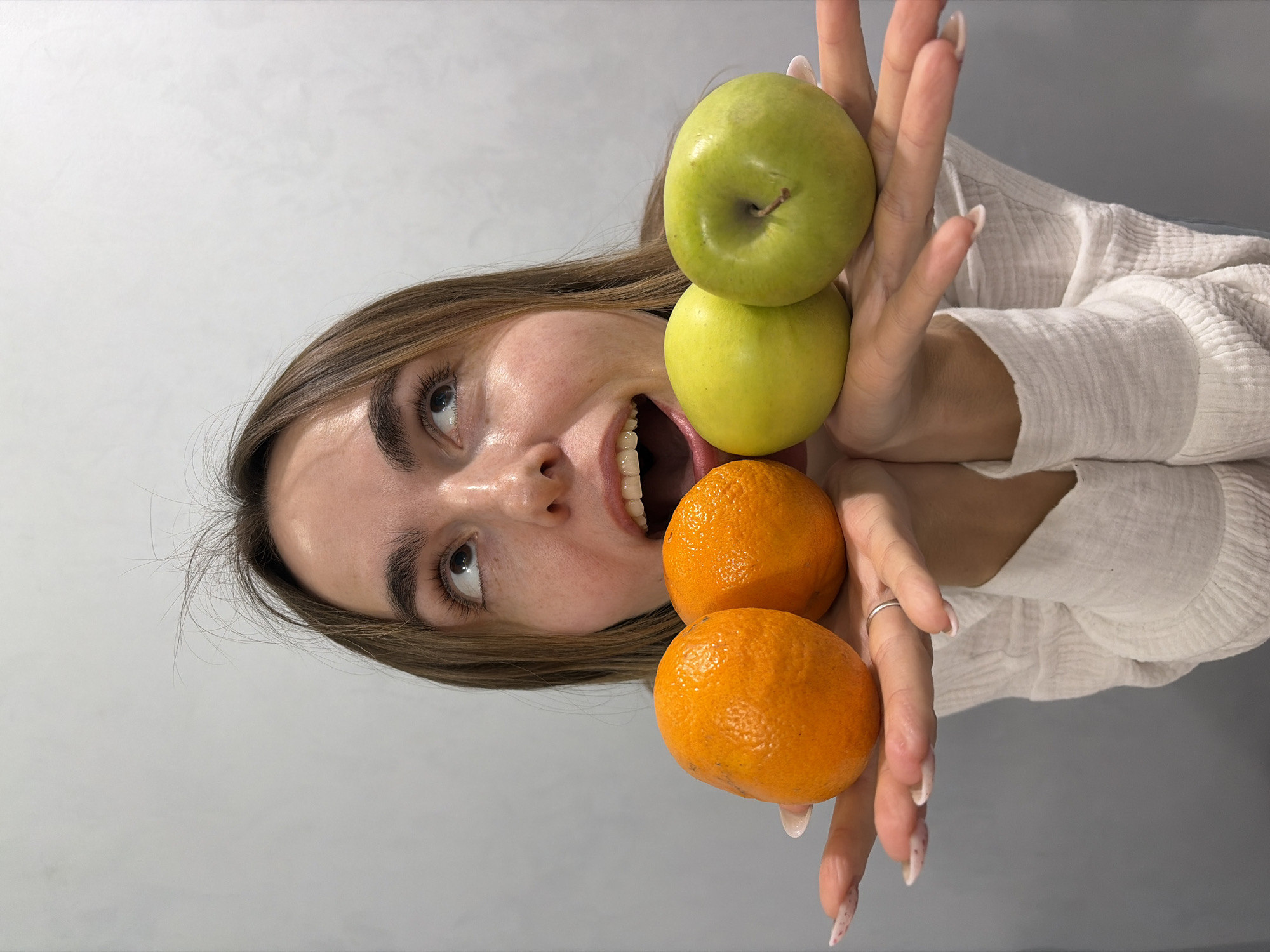 A light-skinned woman with light brown hair is holding two tangerines and two apples (one green and one yellow) in her hands. Her mouth is open, and she is looking up and to the left. She is wearing a white shirt. The background is a light gray wall. There is no indication of the location being Neftekamsk, Russia, or any other specific city.