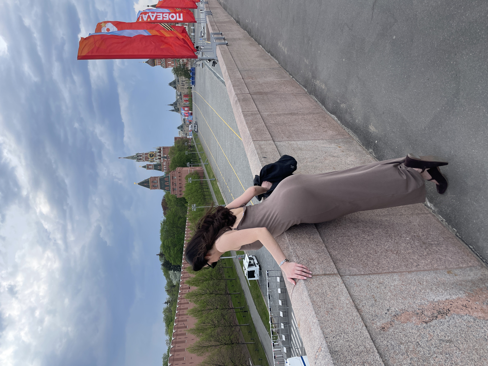 A woman in a brown dress is leaning on a stone wall, with her hands on the wall and her body stretched out. She is wearing sunglasses, and a black purse is slung over her shoulder. Behind her is the Red Square in Moscow, Russia, featuring landmarks such as the Kremlin and St. Basil's Cathedral. Red flags are displayed along the square. The sky is cloudy.